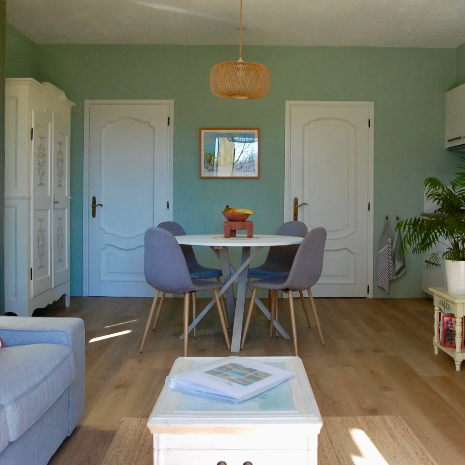 Woonkamer met bank en kussens, ronde eettafel met vier stoelen, witte kast, lichte planten, fles wijn op bijzettafel, grote raam met uitzicht, decoratieve lamp en muurkunst.