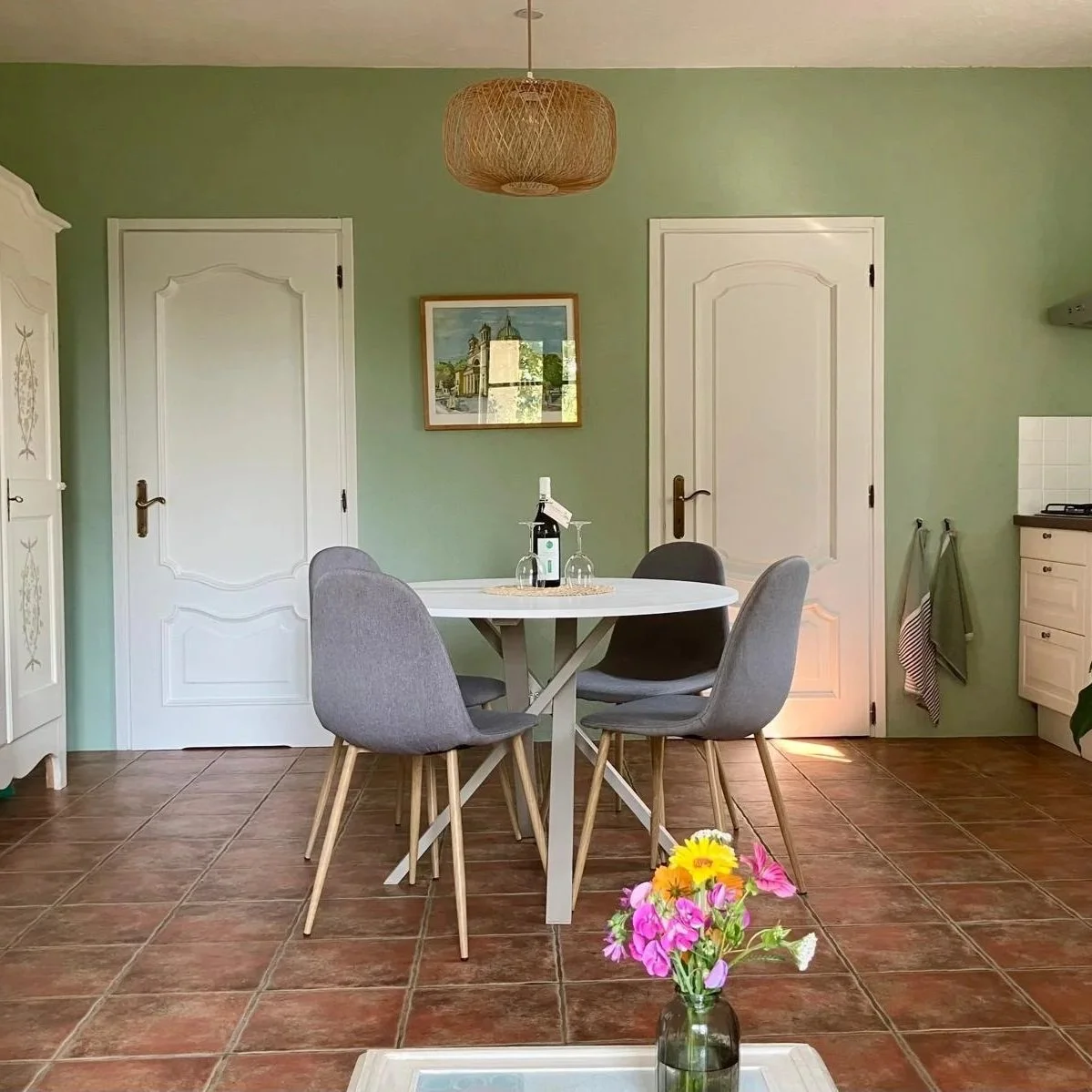 Woonkamer met crèmekleurige bank en kussens, ronde eettafel met vier stoelen, witte kast, lichte planten, fles wijn op bijzettafel, grote raam met uitzicht, decoratieve lamp en muurkunst.