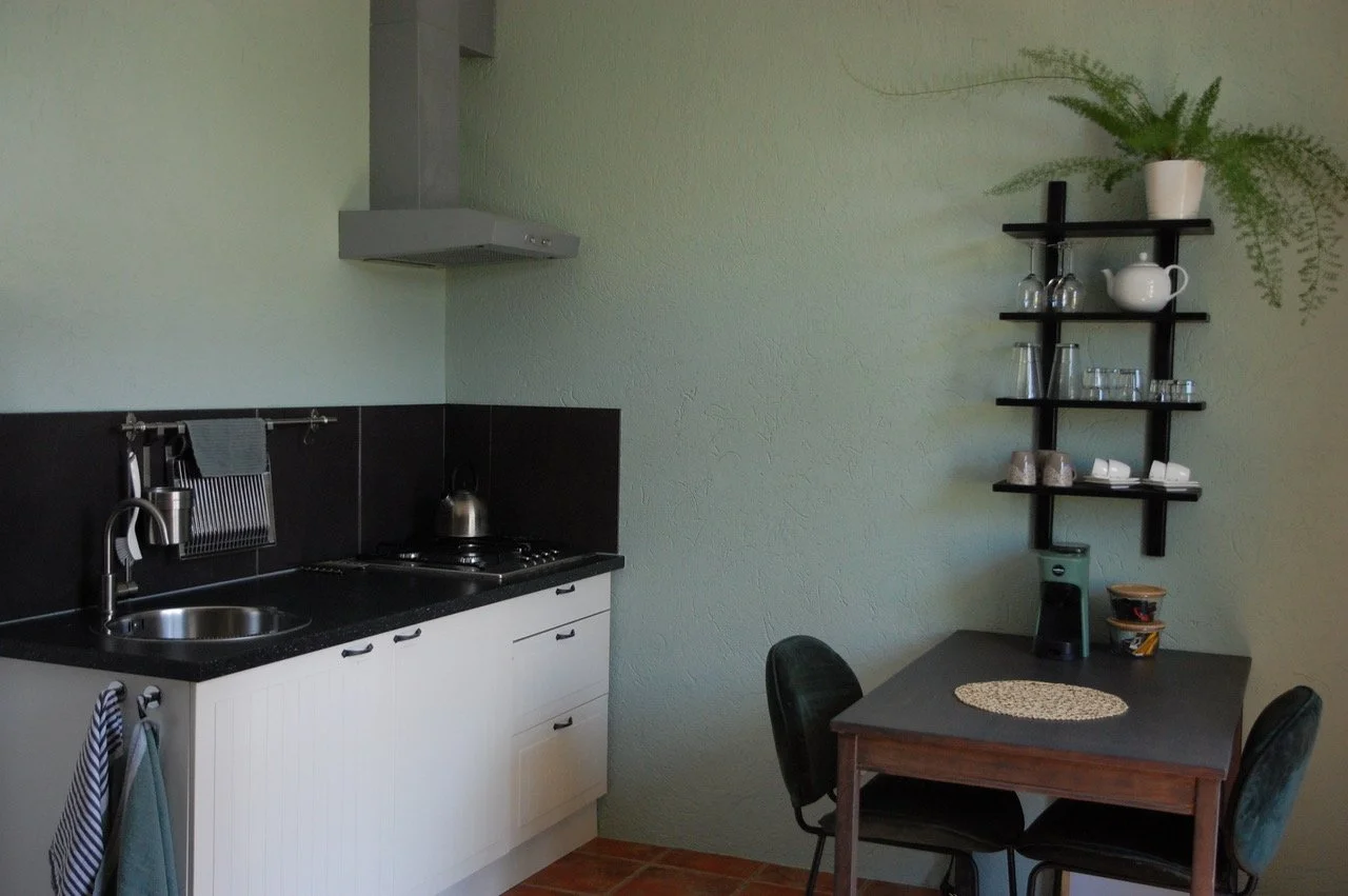 Kleine keuken met wastafel, fornuis, afzuigkap en zwarte kasten. Aan de muur hangt een plank met glazen en mokken. Er is een kleed op een tafel met twee stoelen. Een plant staat op de bovenste plank.