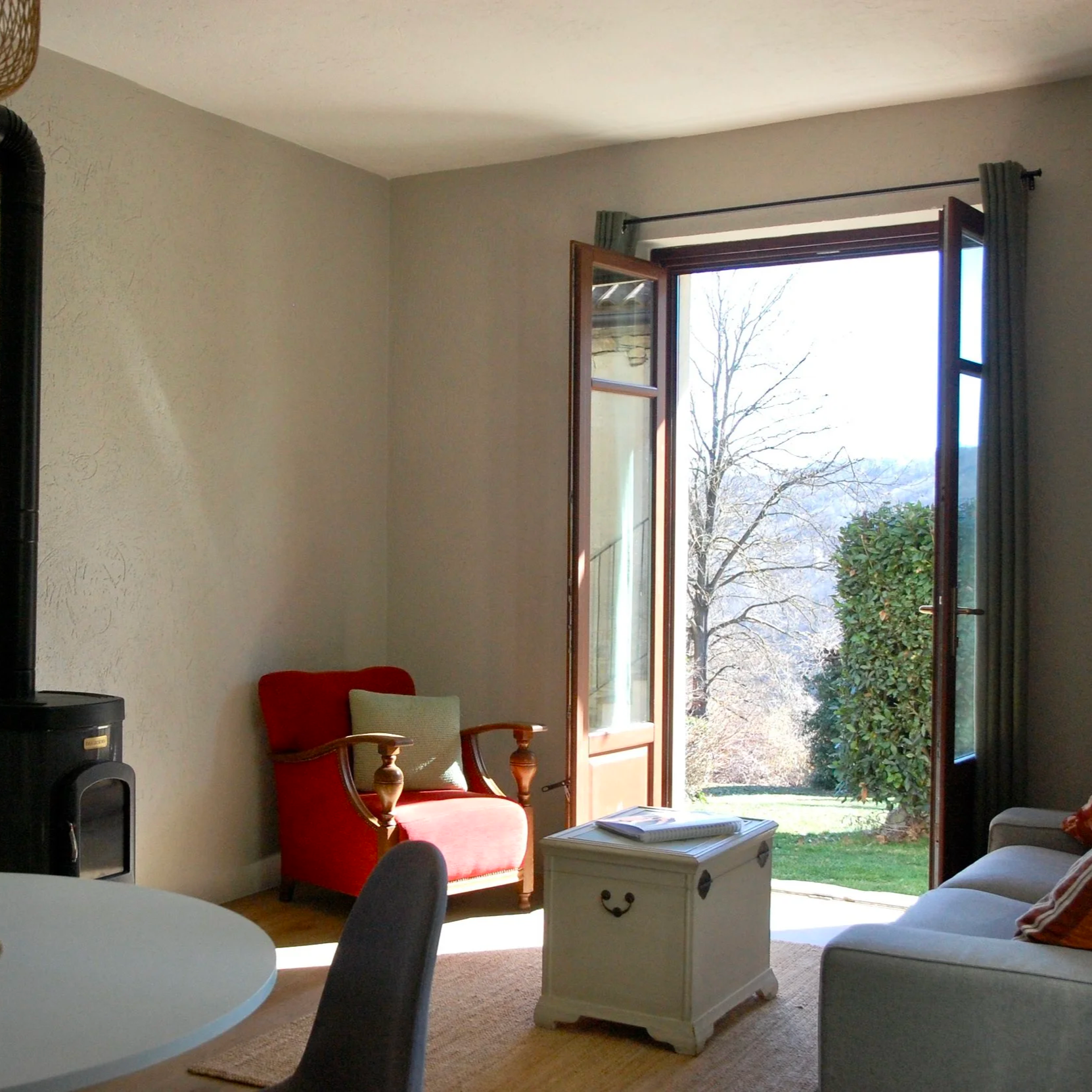 Een gezellige woonkamer met een bank, sierkussens, een houten salontafel, en een fles wijn met glazen. Er is een deur met gordijnen die naar buiten leidt en een raam. Een rode fauteuil staat tegenover een zwarte staande lamp.