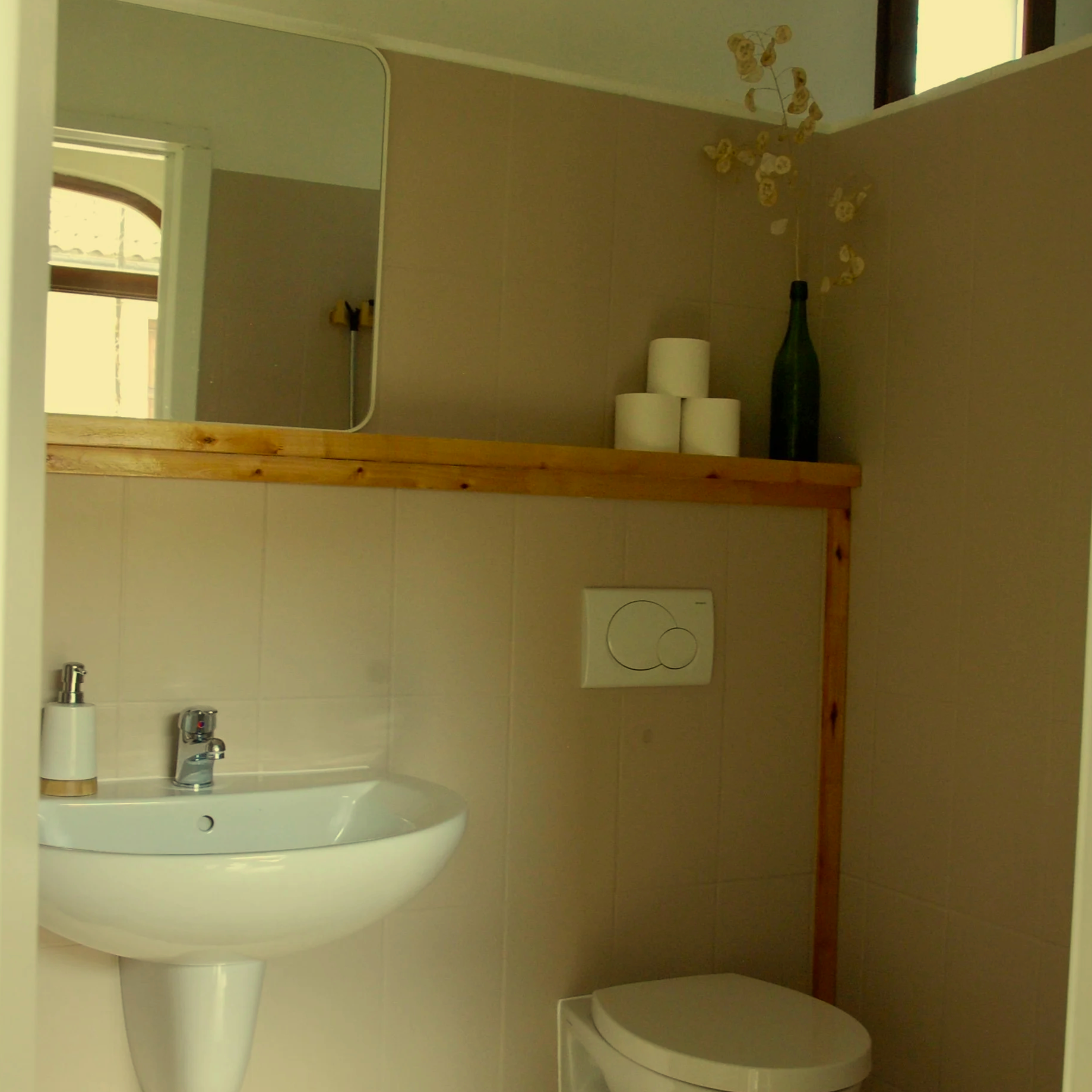 Kleine badkamer met witte wastafel en spiegel er boven. Er is een zeeppompje op de wastafel en een gedeeltelijk zichtbare handdoek.