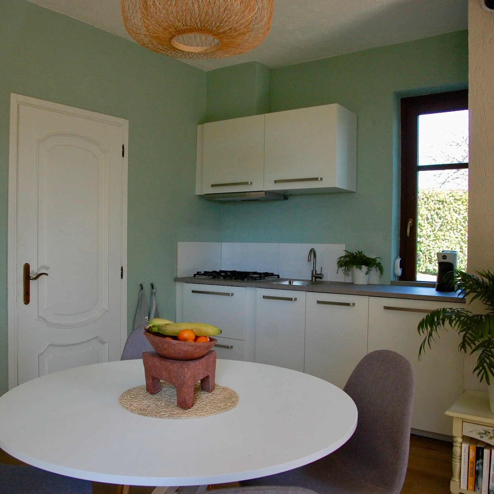 Een moderne keuken met een ronde witte eettafel en vier grijze stoelen. Op de tafel staat een schaal met fruit. De keuken heeft lichtgekleurde kasten, een zwarte kookplaat, en groene muren. Er hangt een gevlochten lamp aan het plafond.