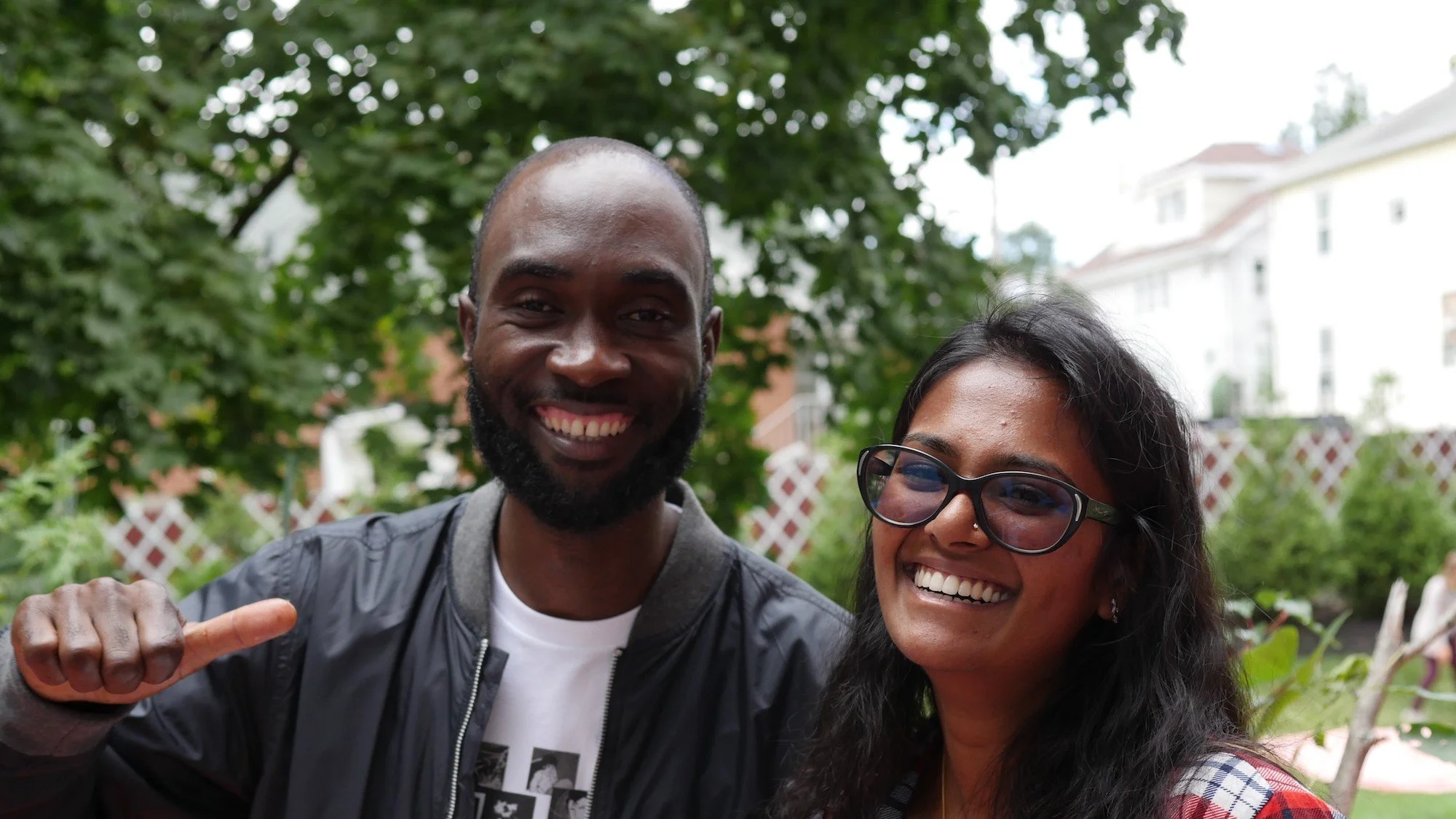 A smiling man with a beard and a bald head giving a thumbs-up, standing beside a woman with glasses and dark hair, both outdoors with green trees and residential buildings in the background.