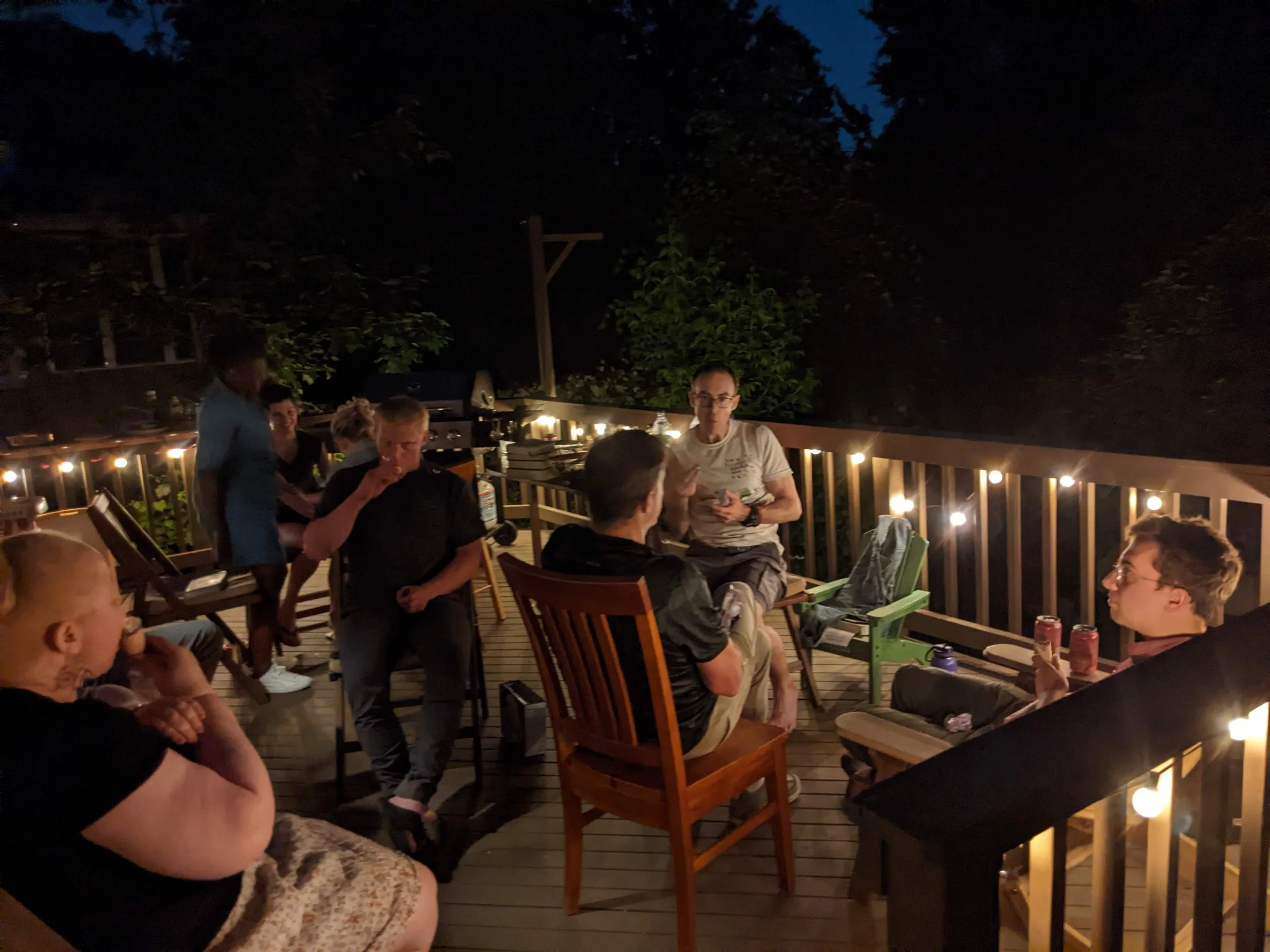 Group of people sitting and standing on a wooden deck at night, illuminated by string lights, with trees and a dark sky in the background.