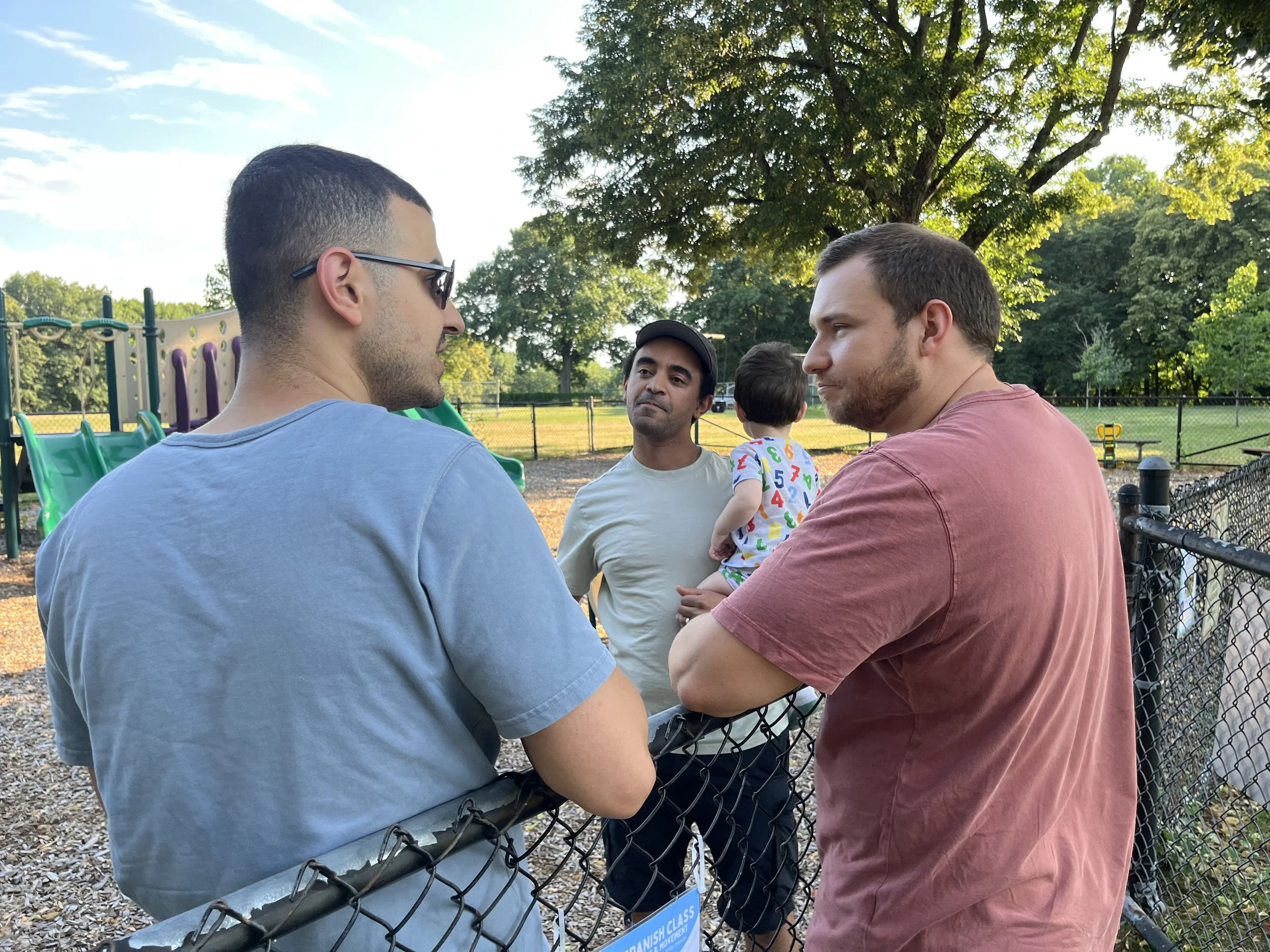 Two men are having a conversation at a park near a playground, with a small child standing on a woman’s lap. The men are facing each other, and there are trees and a fence in the background.