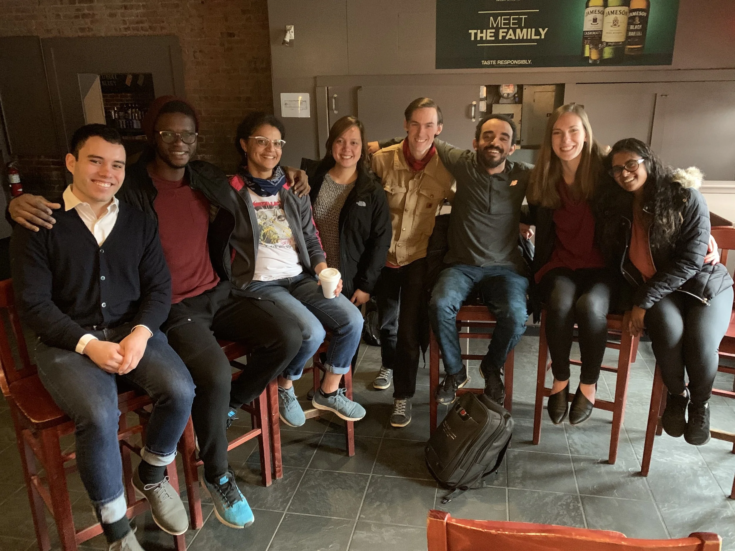 A group of eight diverse young adults sitting on barstools in a restaurant or bar, smiling at the camera, with two of them holding coffee cups. They are sitting closely together with arms around each other. The setting has brick and gray walls with a sign above that says 'Meet the Family' and bottles of Jameson Irish whiskey on display.