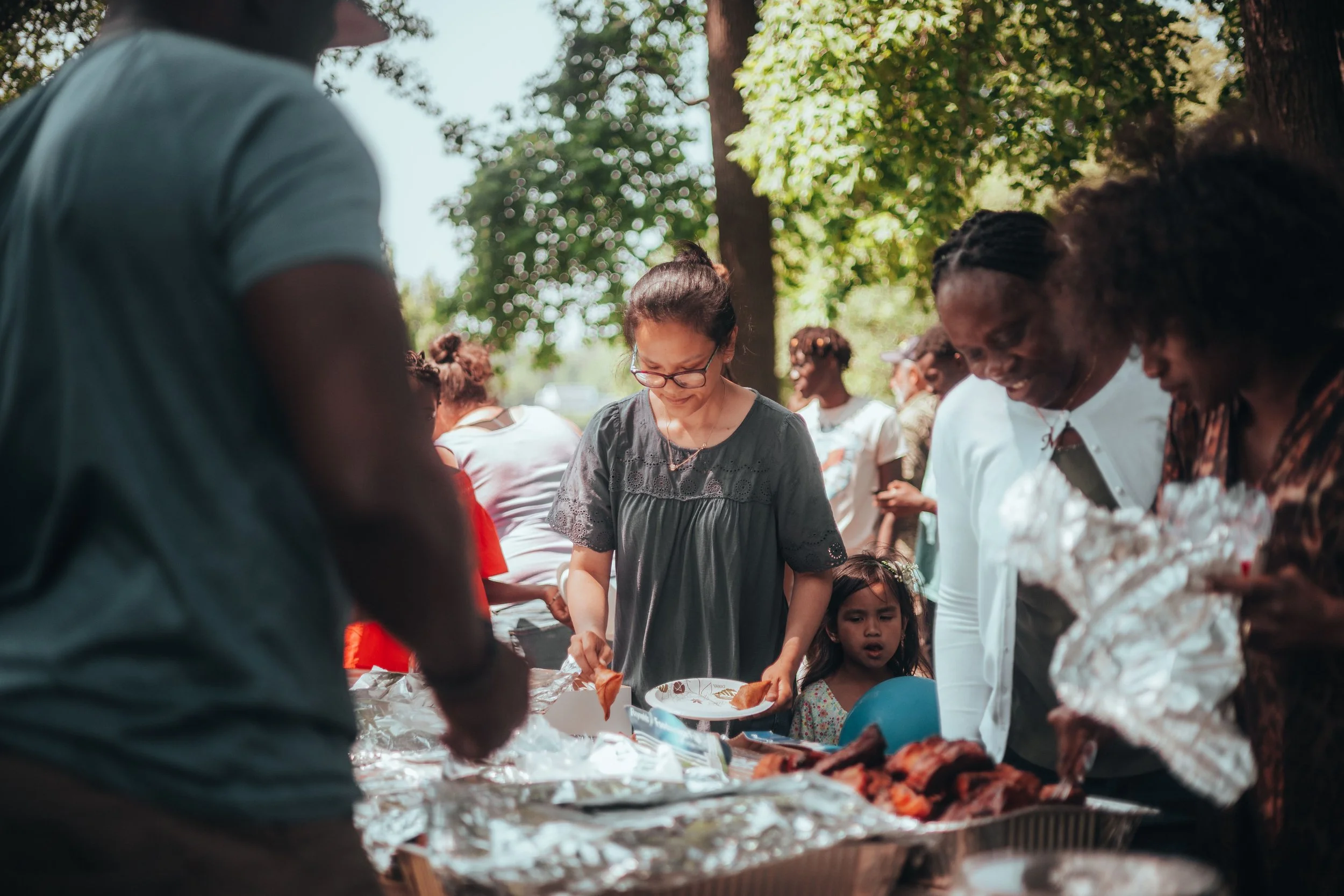 A diverse group of people gathered outdoors around a table with food, enjoying a communal event in a park-like setting with trees and sunlight.