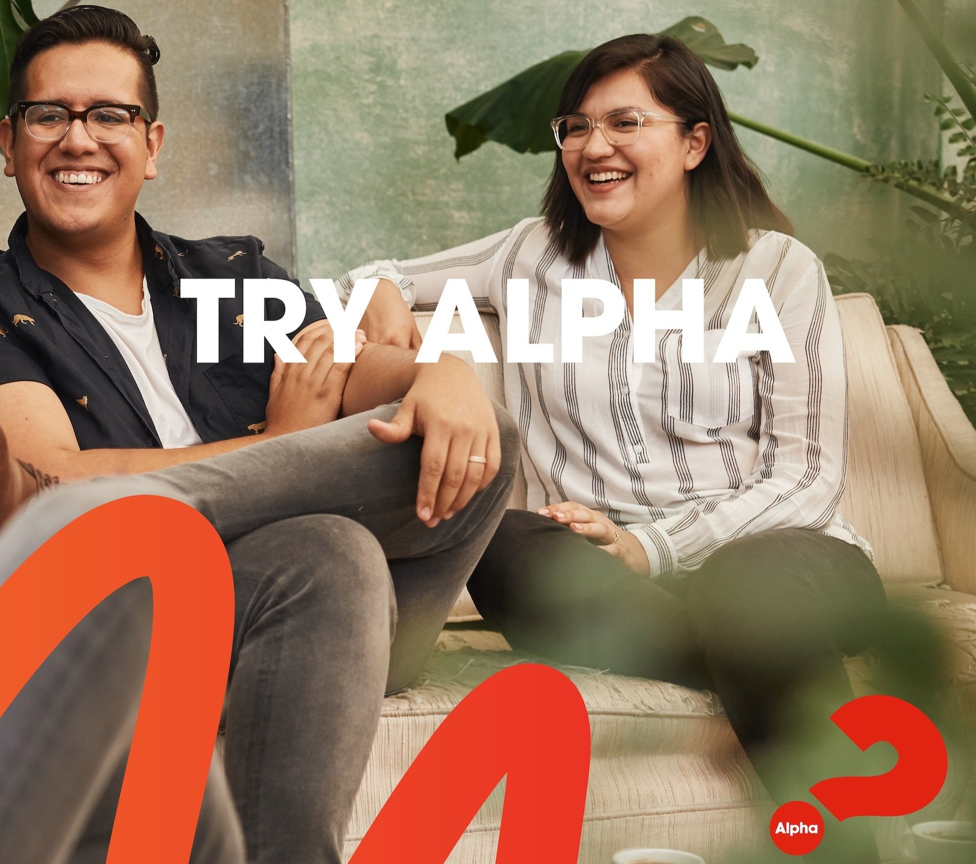 Two people sitting on a couch, smiling and laughing. The man on the left has glasses and dark hair, wearing a black shirt with a pattern. The woman on the right has glasses and dark hair, wearing a white shirt with thin black stripes. There is a large green plant with broad leaves in the background. The image has red, orange, and green graphic elements overlaid, with the word "Alpha" in a red circle and the text "TRY ALPHA" at the bottom.