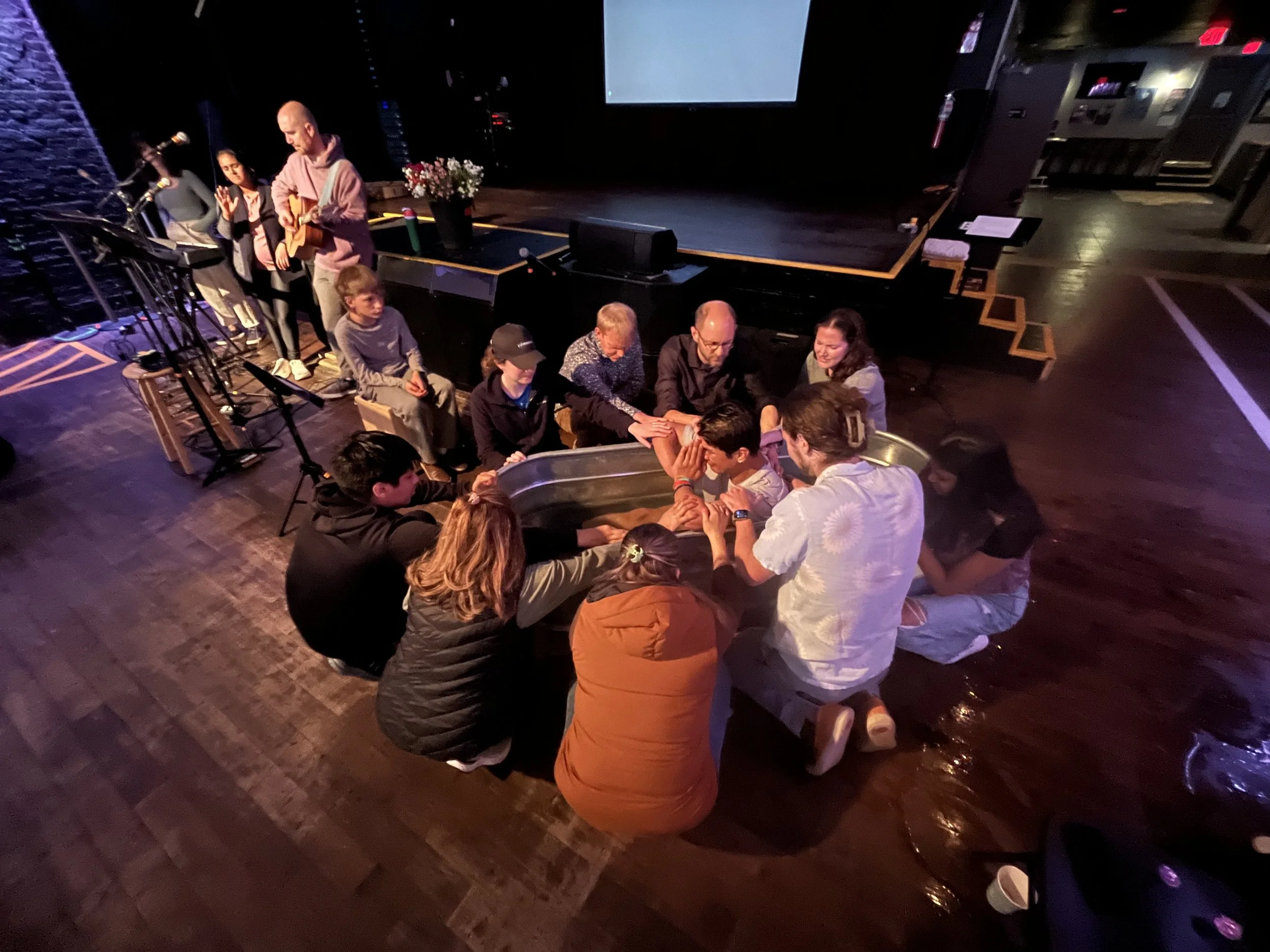 A group of people seated in a circle, touching hands in a prayer or meditation practice, in a dimly lit indoor space with a stage, flowers, and a large screen overhead.