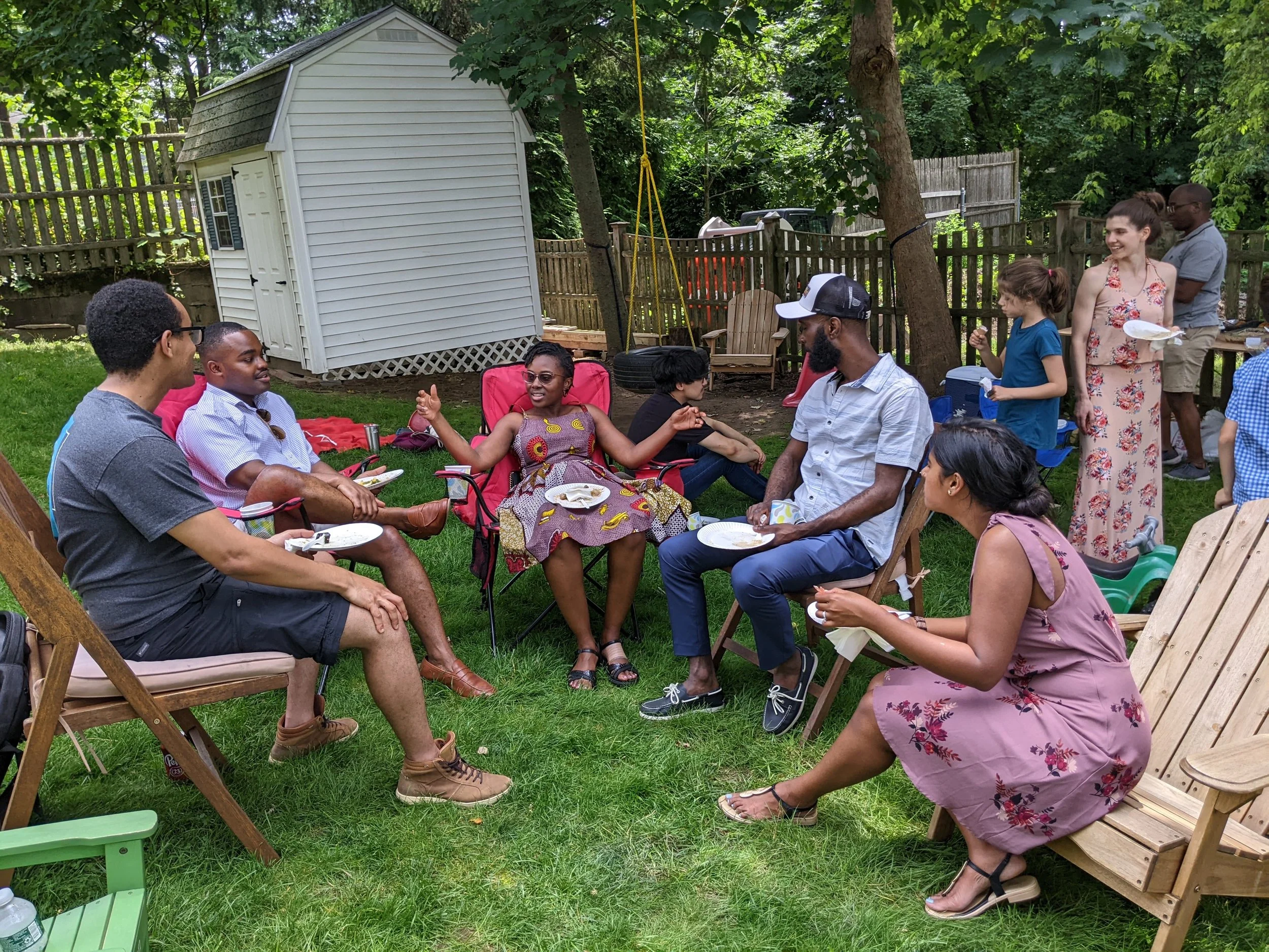 A diverse group of people gathered in a backyard for a social event, sitting on outdoor chairs, some eating, and engaging in conversation. There are trees, a wooden fence, and a small white shed in the background.