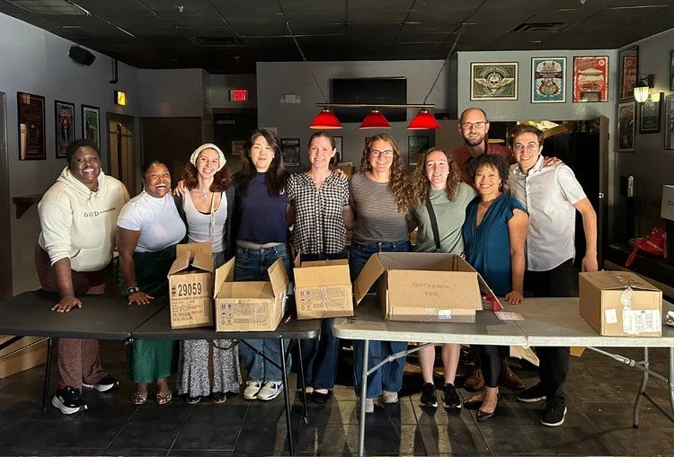 Group of ten people standing behind a long table with empty cardboard boxes inside, in a room with framed pictures on the wall.