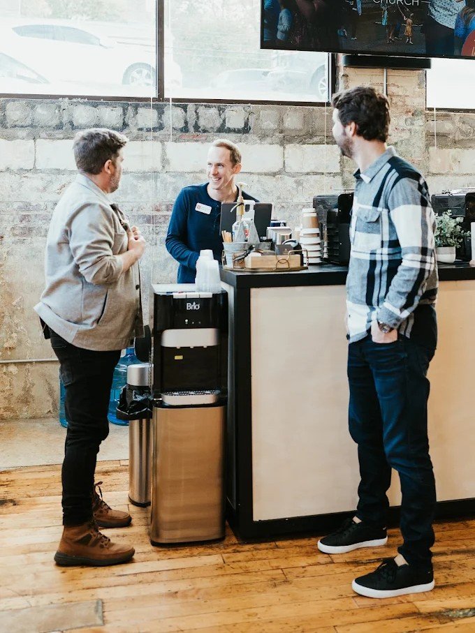 Three men having a conversation at a coffee shop counter with a barista behind it, inside a modern cafe.