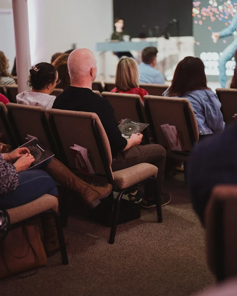 Audience seated in rows at an indoor event watching a performer on stage, with some audience members holding programs or brochures.