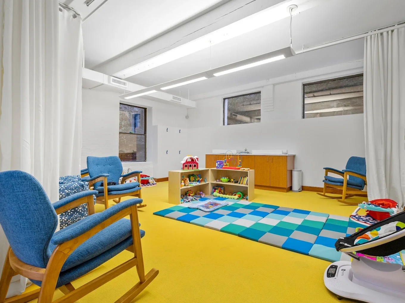 Empty children's playroom with blue chairs, a colorful foam mat, toys on wooden shelves, and large windows.