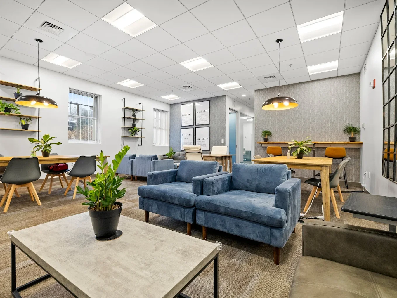 Modern office lounge with blue velvet sofas, wooden tables, chairs, and shelves with plants, bright lighting, and framed wall art.