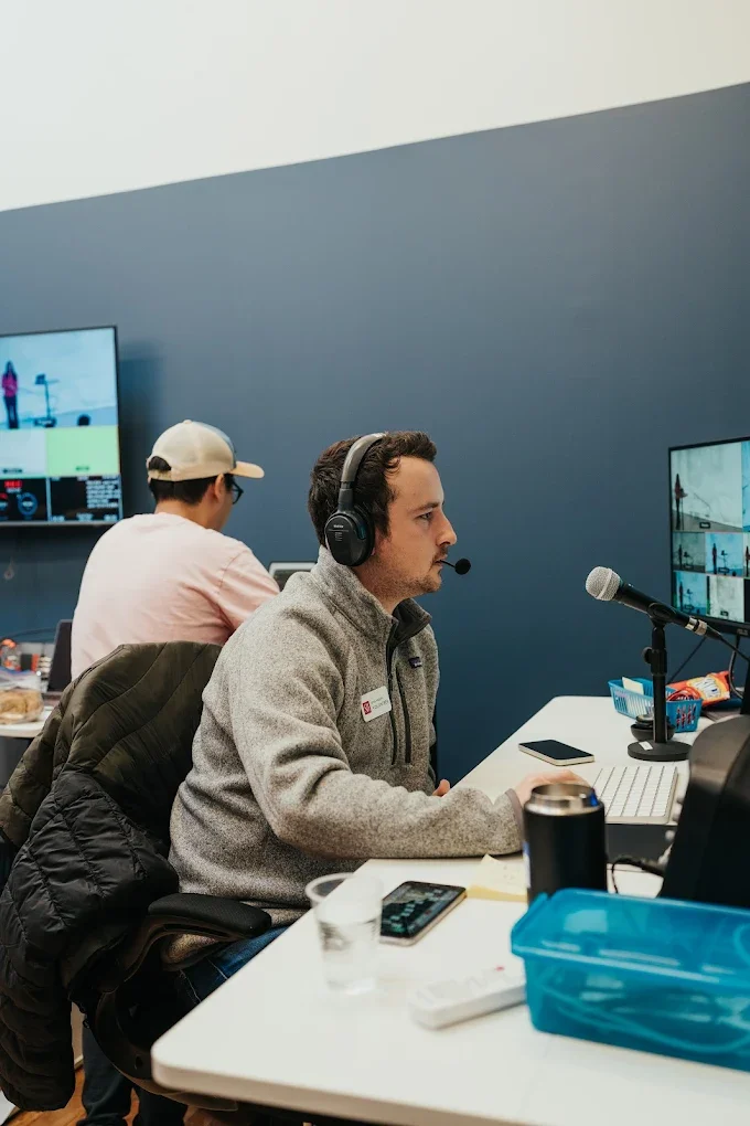 A man with headphones and a microphone working at a desk with a computer, and another person working at a separate desk in the background.