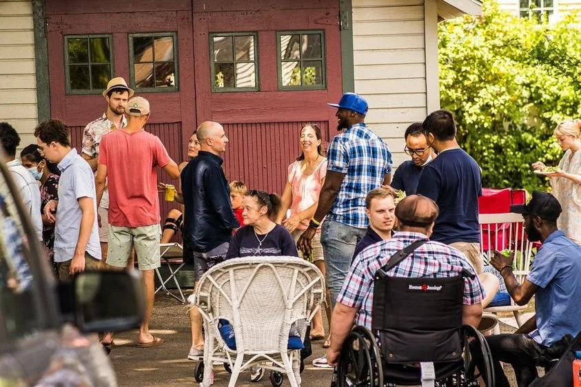 A diverse group of people enjoying a gathering outdoors in front of a house with a red garage door and white siding. Some are standing, some are seated, and they are chatting and smiling.