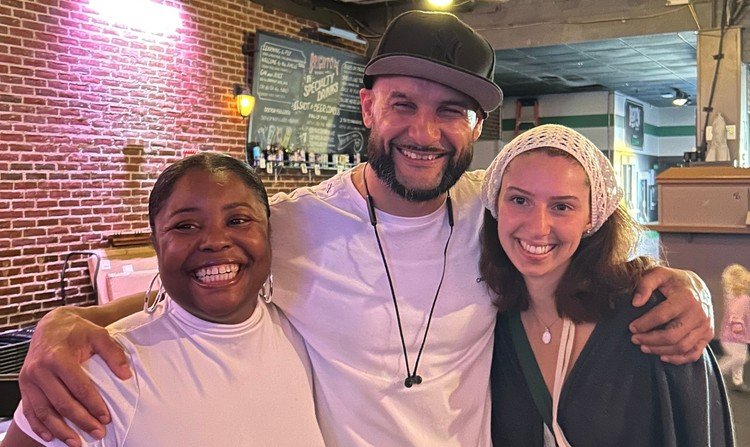 Three people smiling and hugging inside a restaurant or bar with a brick wall and chalkboard menu in the background.