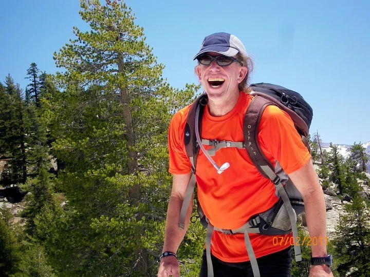 A man in orange hiking clothes, wearing a baseball cap and sunglasses, smiling and laughing while hiking through a forested area.