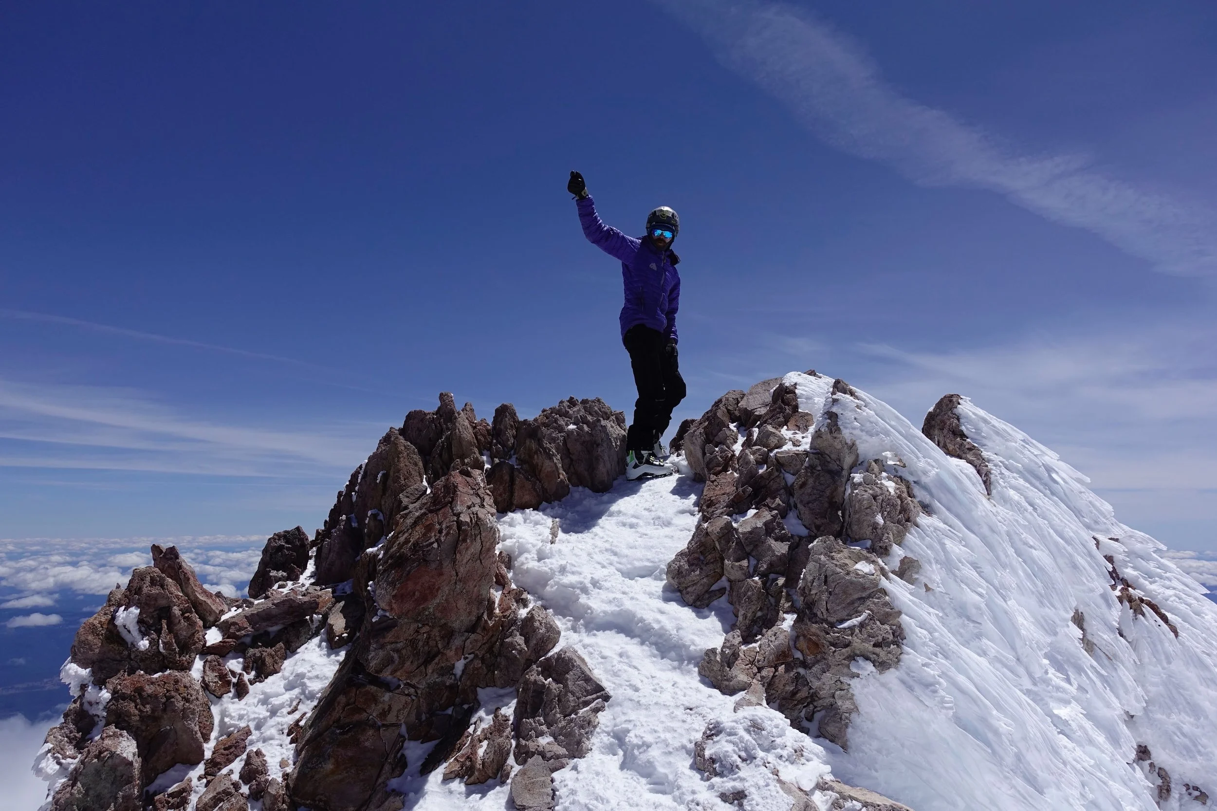 Person in winter clothes and climbing helmet standing atop the snowy summit of Mount Shasta