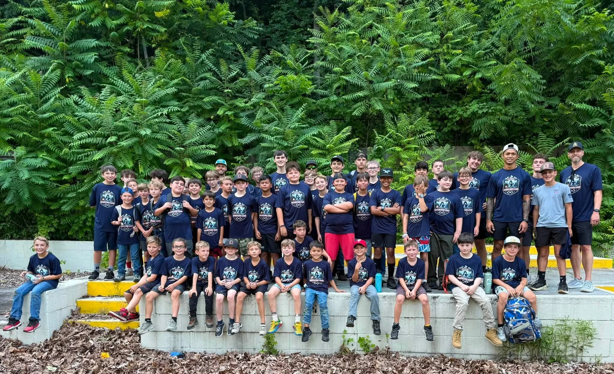 Group of children and adults posing outdoors in front of a lush green, fern-filled wooded background.