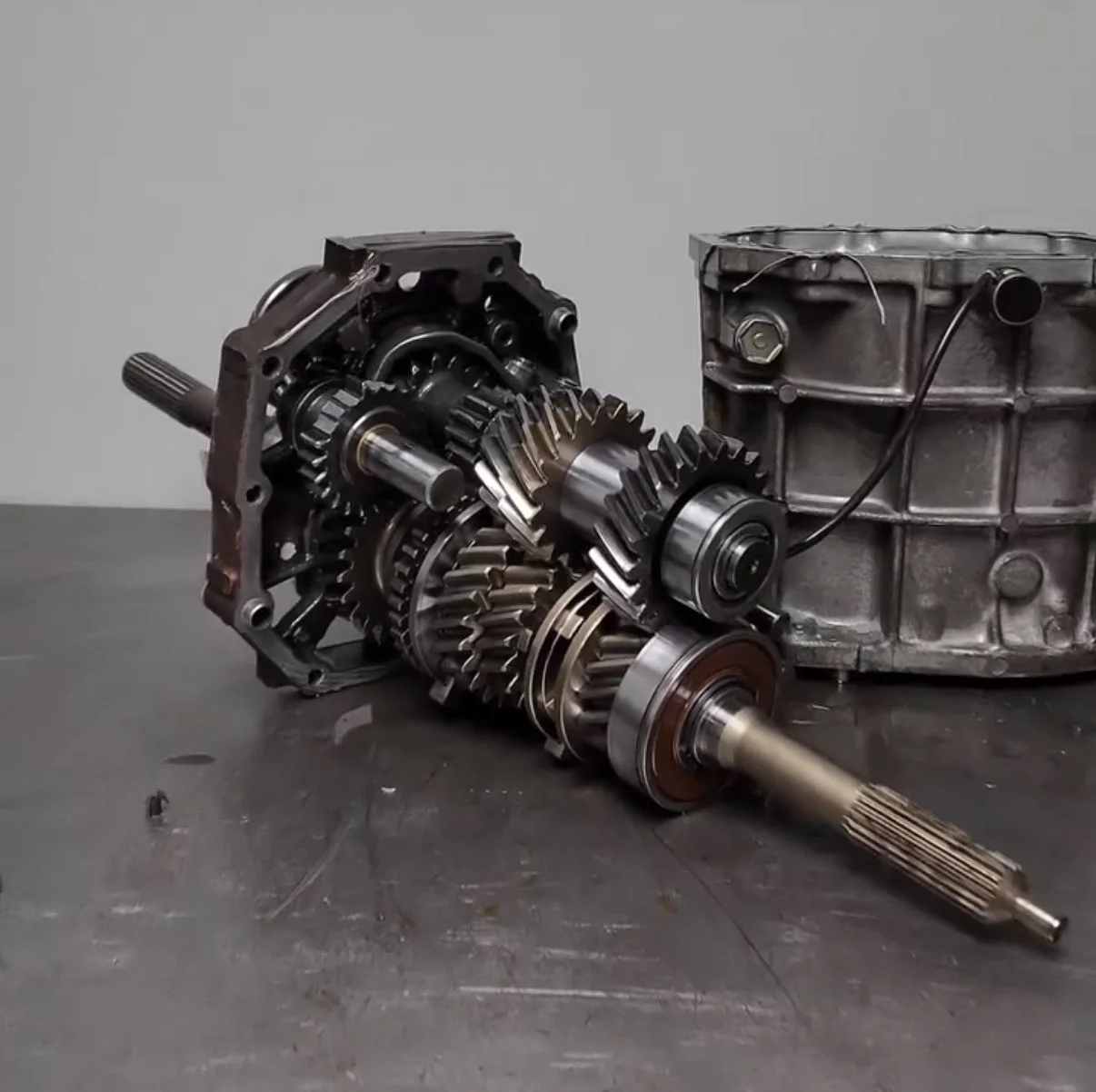 Disassembled gearbox with gears and shafts next to a metallic transmission case on a gray work surface.