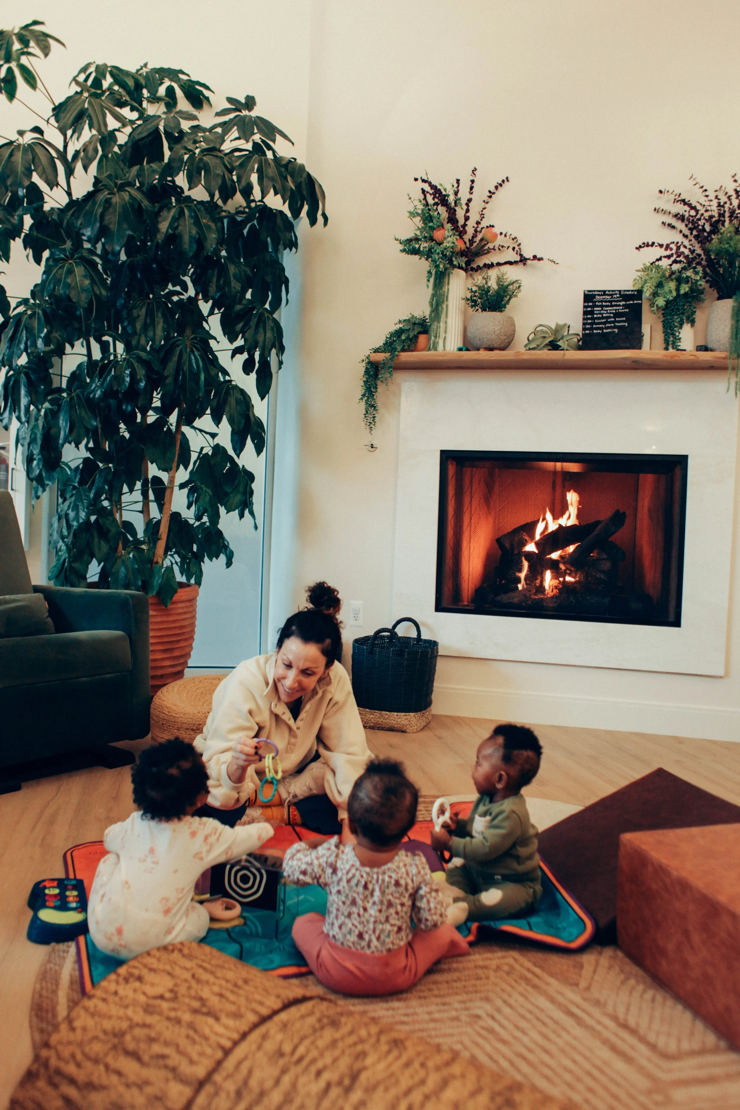 A woman standing on a rug in front of a fireplace with a fire, surrounded by children seated in chairs, in a cozy room with a large plant and decorative shelves.
