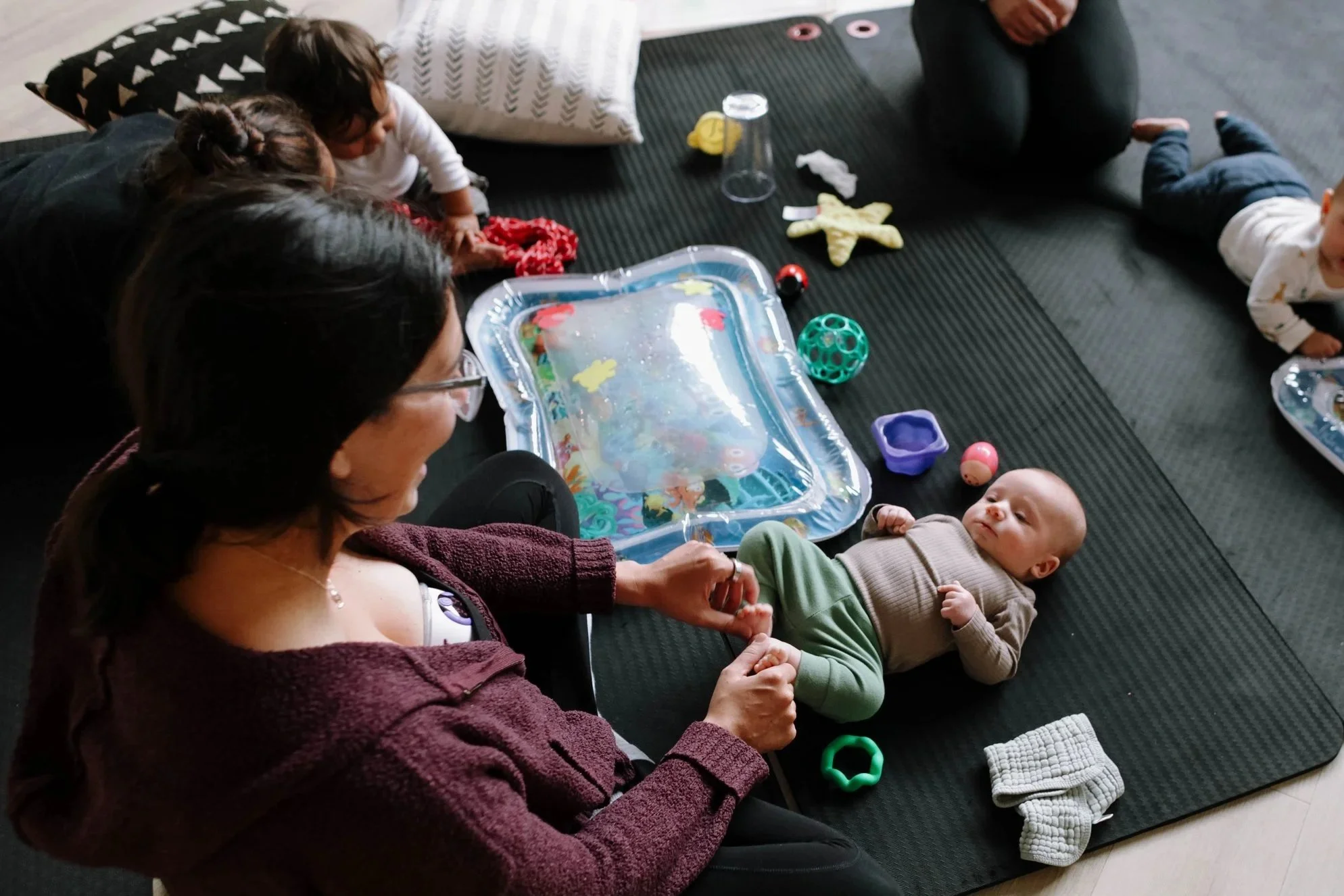 A woman leading a yoga class in a studio, sitting cross-legged on a yoga mat and stretching her arms overhead, with other women in the background sitting on mats and stretching.