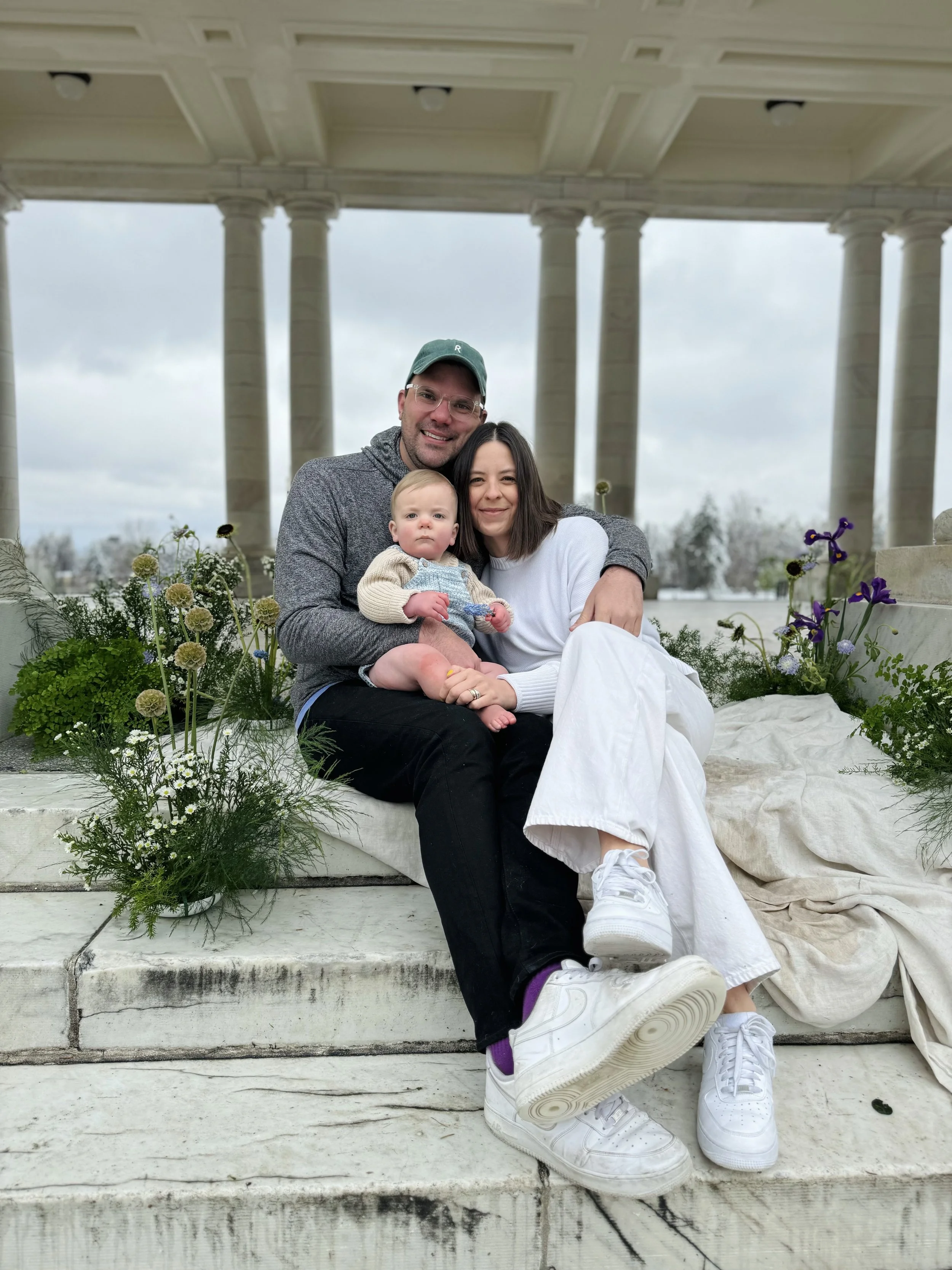 A family of three sitting on white marble steps outdoors under a colonnade, surrounded by flower arrangements. The man, woman, and child are smiling, with the man holding the baby, and the woman leaning in close.