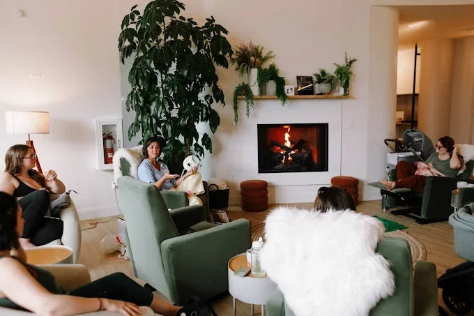 A woman standing on a rug in front of a fireplace with a fire, surrounded by children seated in chairs, in a cozy room with a large plant and decorative shelves.