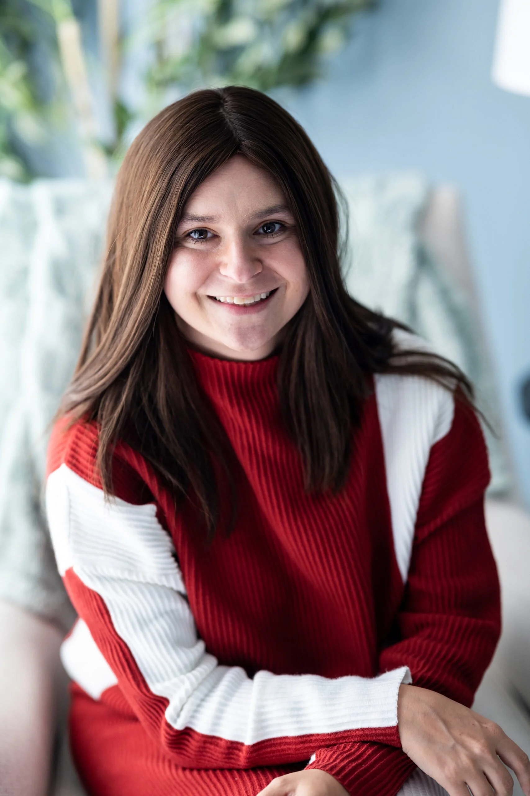 A young woman with long brown hair smiling, wearing a red and white sweater, sitting indoors with a blurred background of greenery.