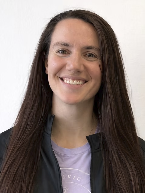 A woman with long brown hair smiling at the camera, wearing a black jacket and a light purple shirt, against a plain white background.