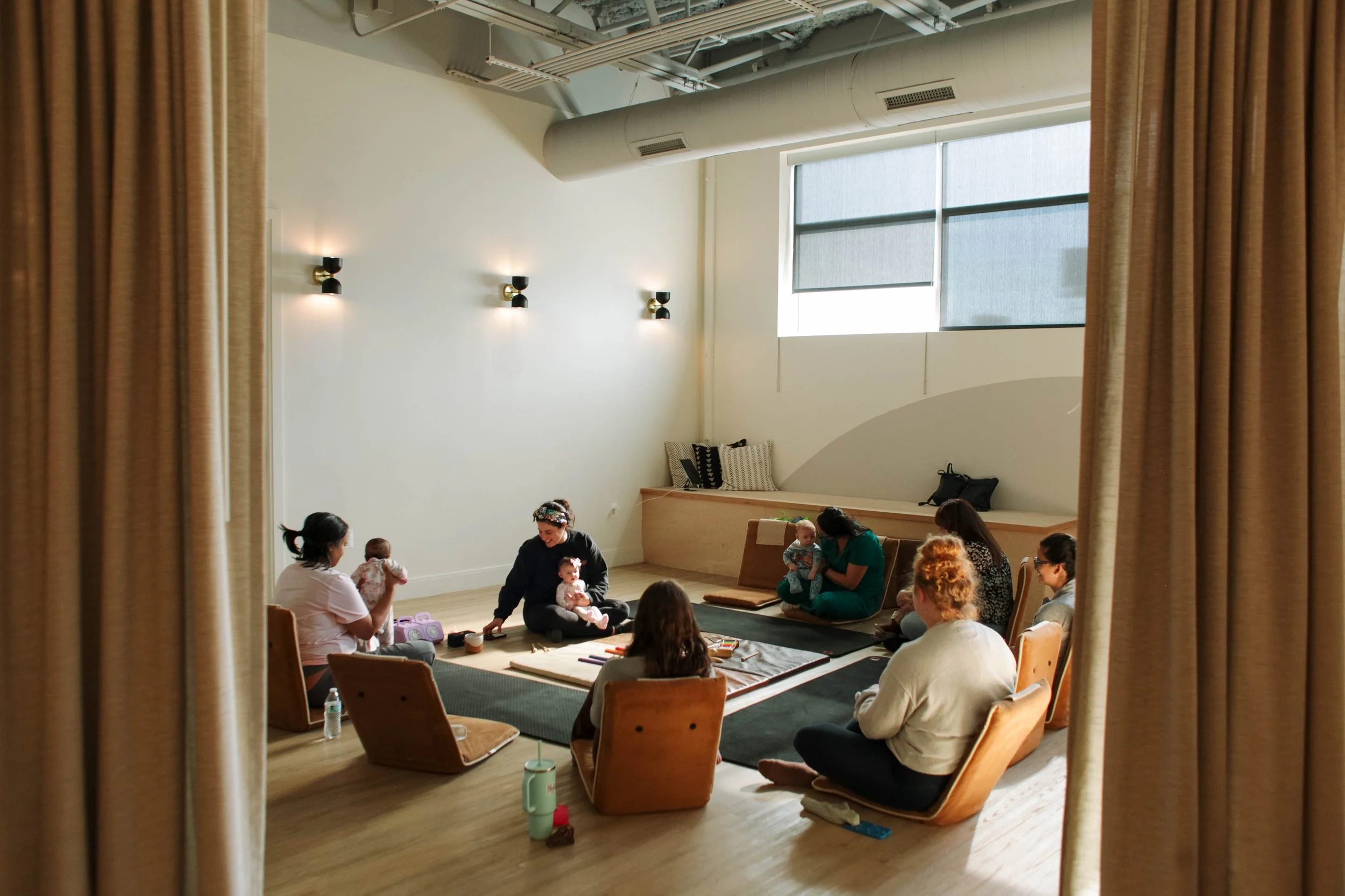 A group of people in a prenatal yoga class, some in child's pose, on yoga mats in a studio, with a yoga instructor sitting at the front.