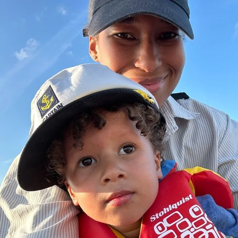 A woman smiling with a young boy, who is wearing a sailor hat and a red life jacket, outdoors with a blue sky in the background.