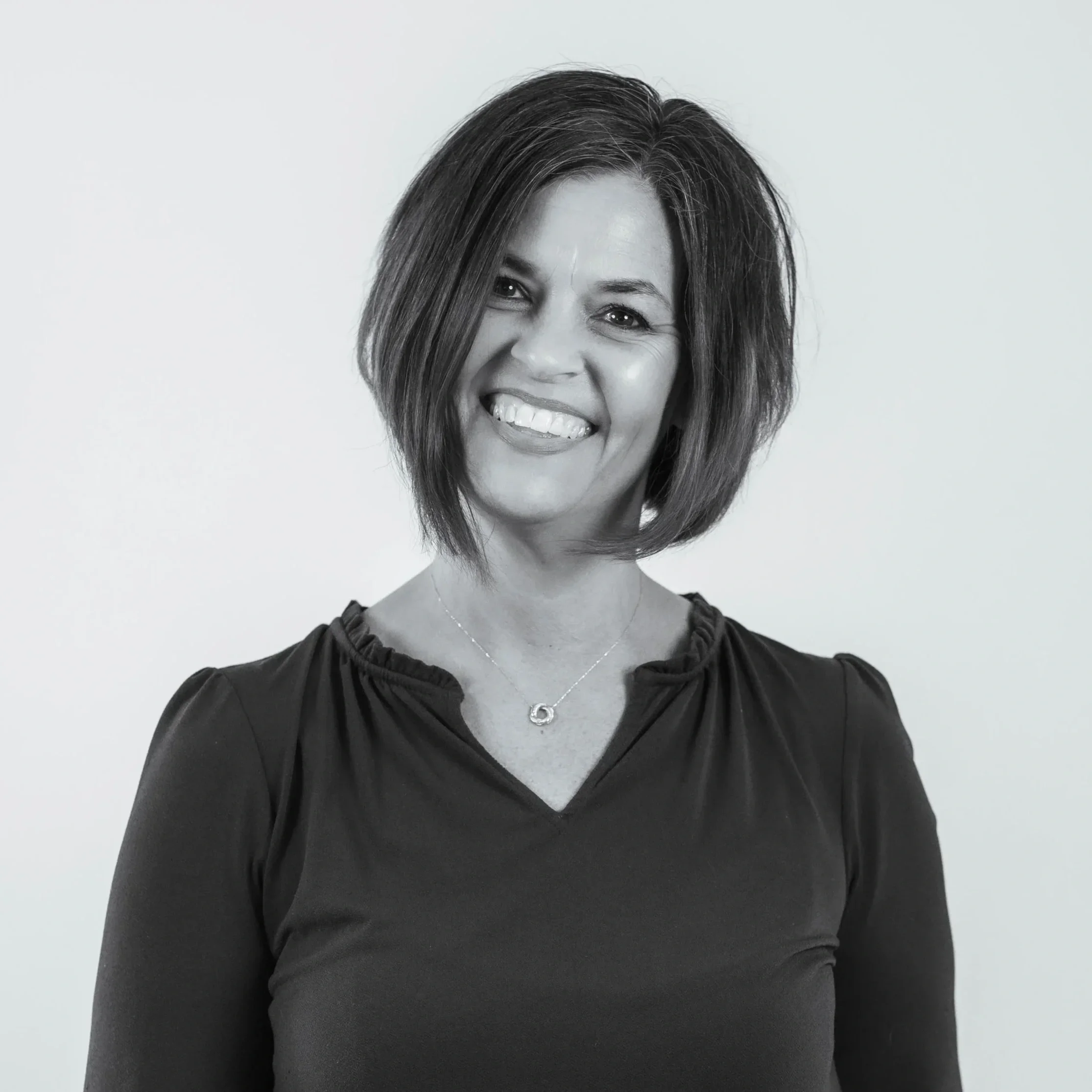 A woman with short, dark, layered hair smiling and showing her teeth. She is wearing a black top with a ruffled neckline and a delicate necklace.