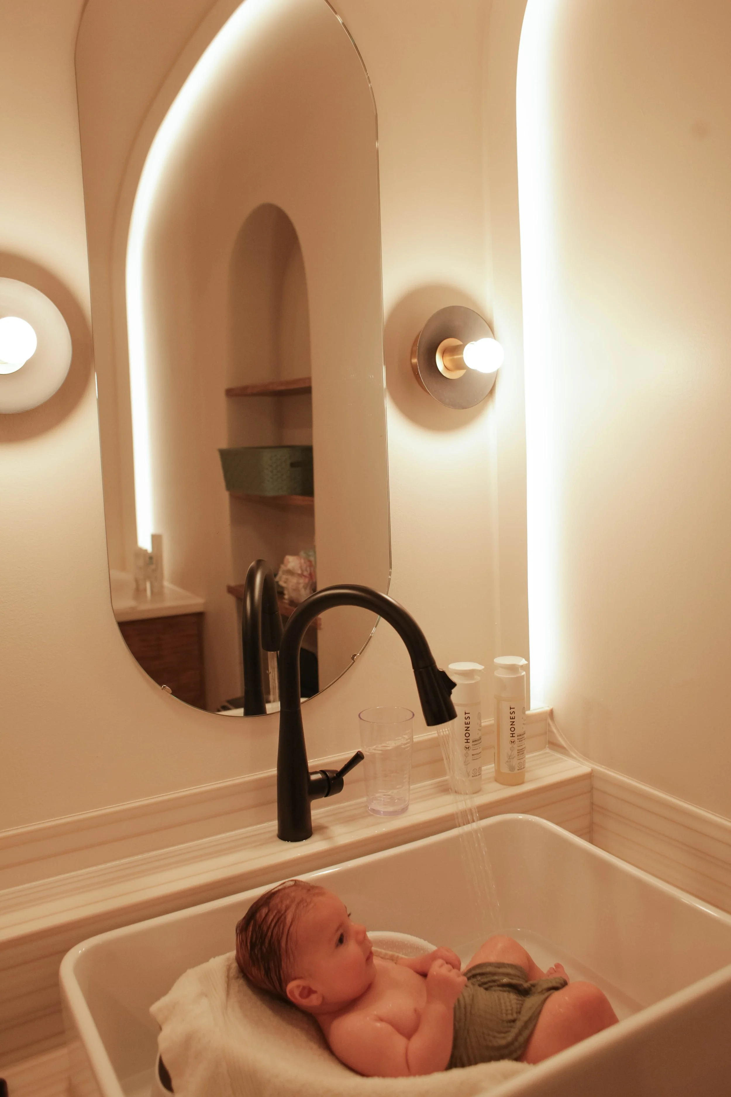 Baby with a hat in a bathtub, being held by an adult, with water running from the faucet.