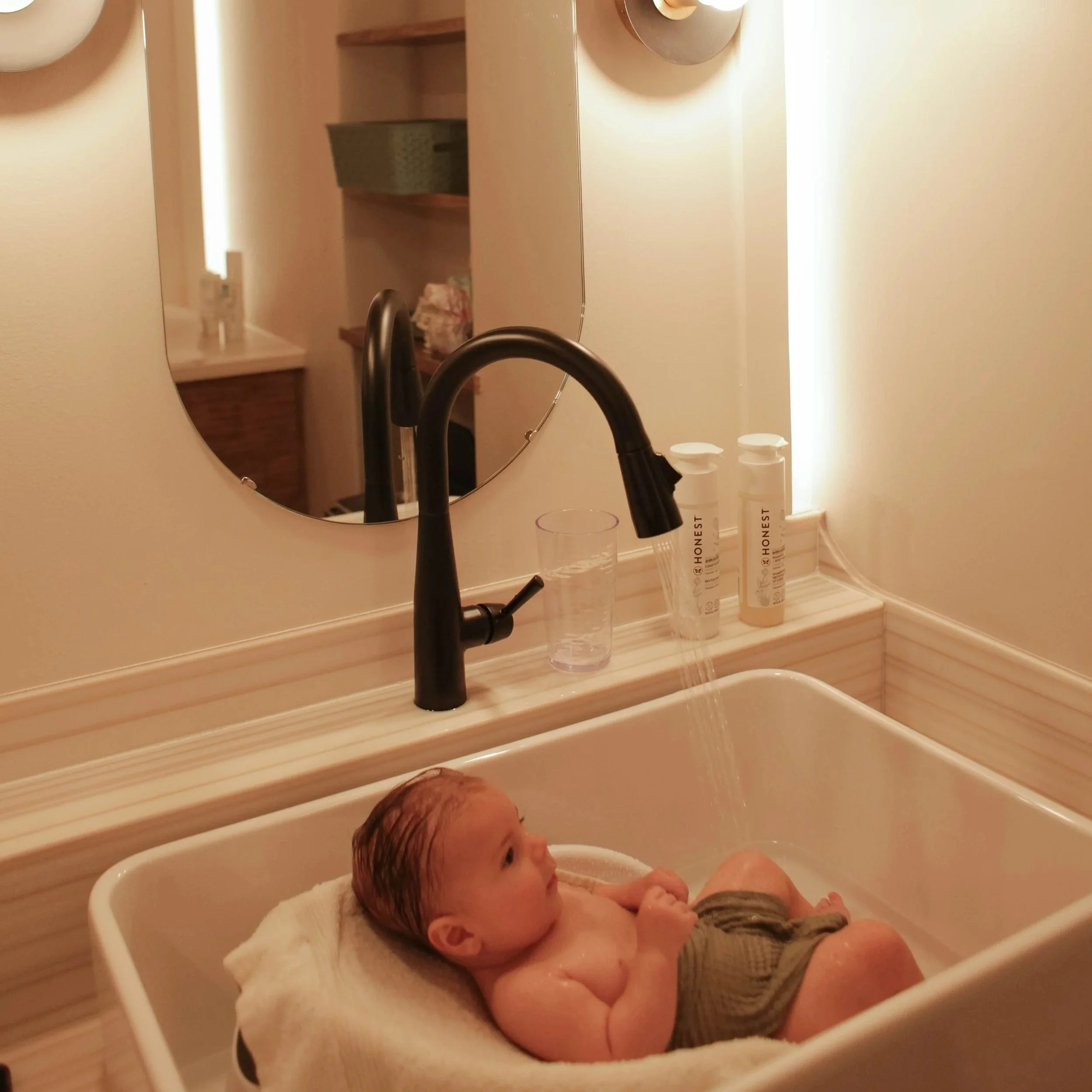 Baby with a hat in a bathtub, being held by an adult, with water running from the faucet.