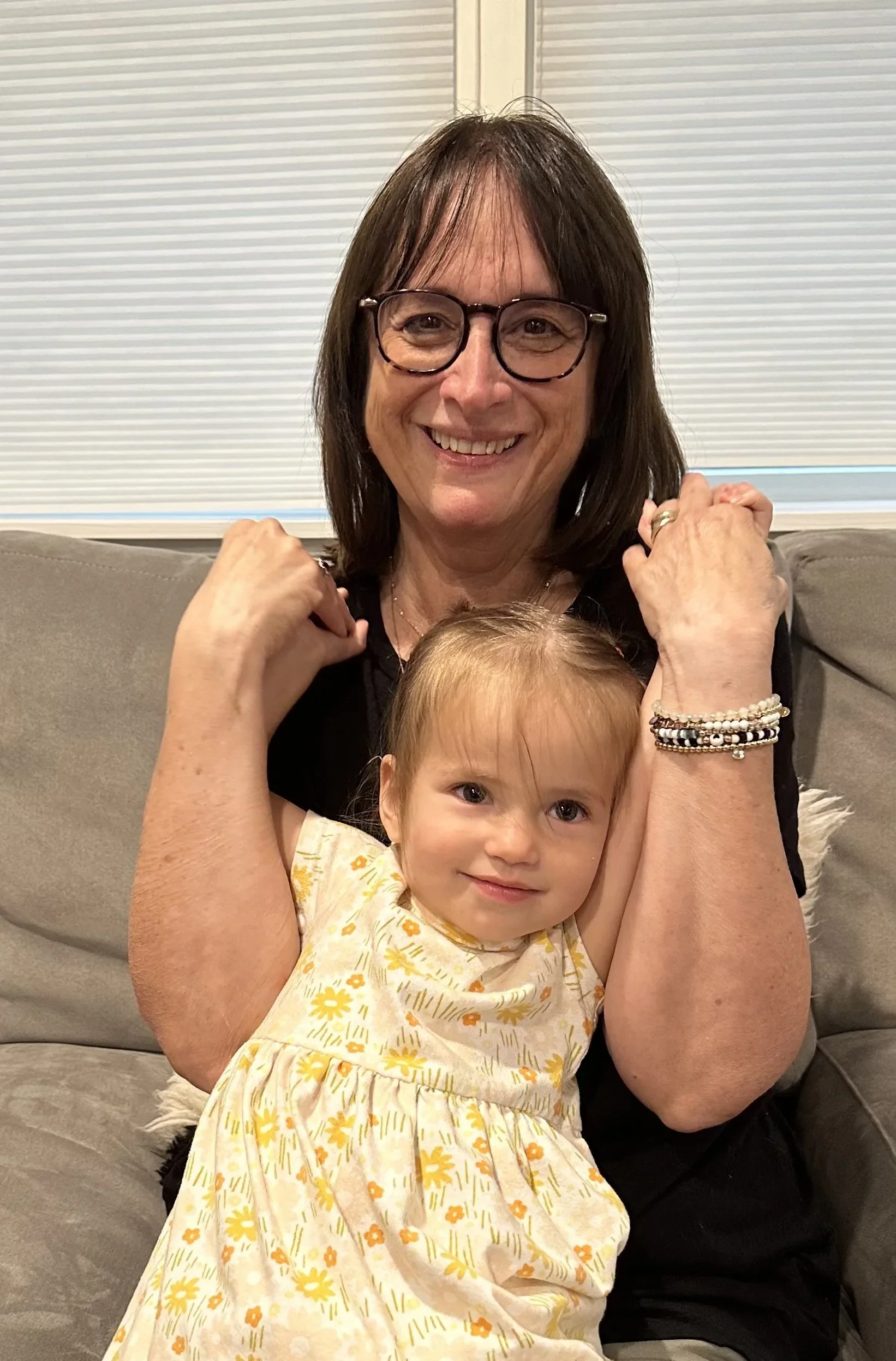 A woman with glasses and a young girl sitting on a gray couch, smiling and hugging at home.