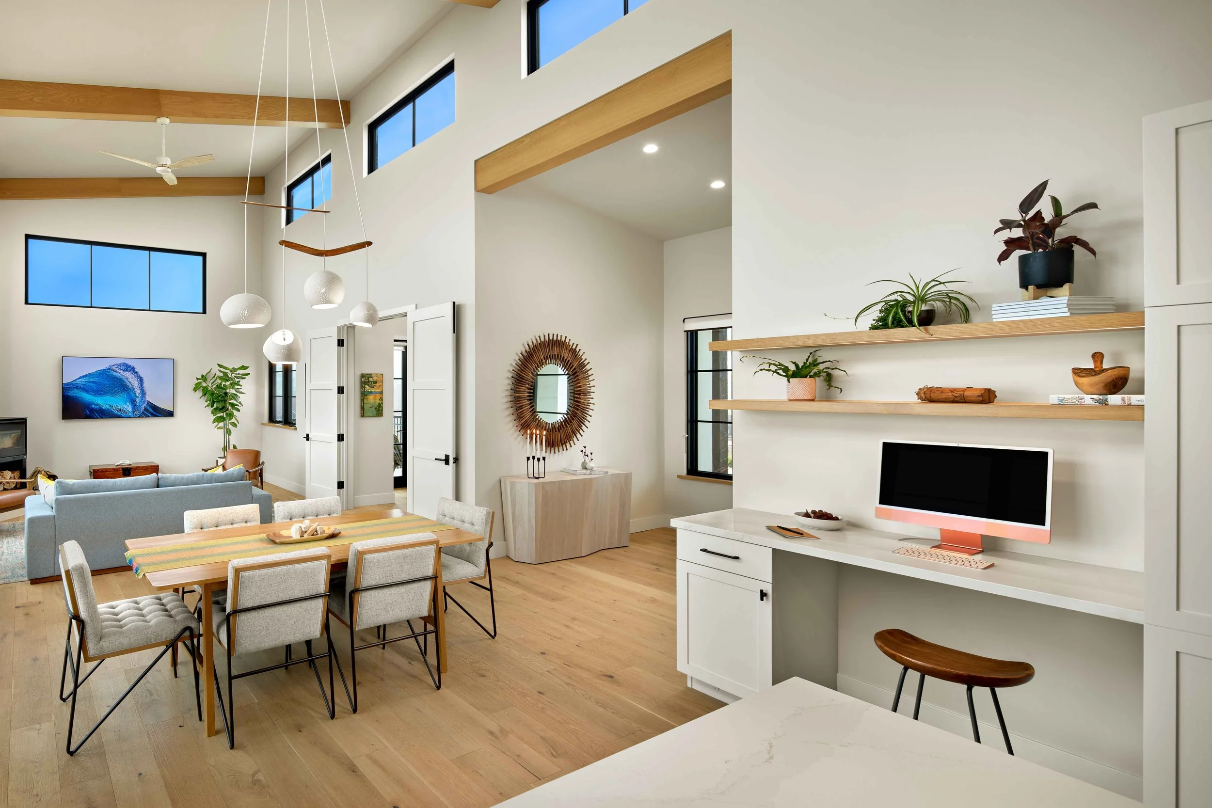 A modern, open-concept living and dining area with high ceilings, large windows, and wooden accents. The space features a dining table with fabric chairs, a TV mounted on a wall, and decorative plants on floating wooden shelves.