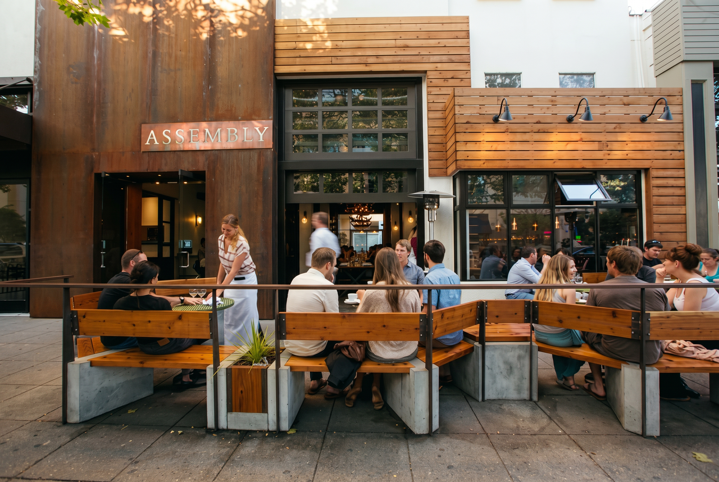 Outdoor patio with wooden seating and people dining at a restaurant called Assembly, with a wooden and metal exterior and large windows.