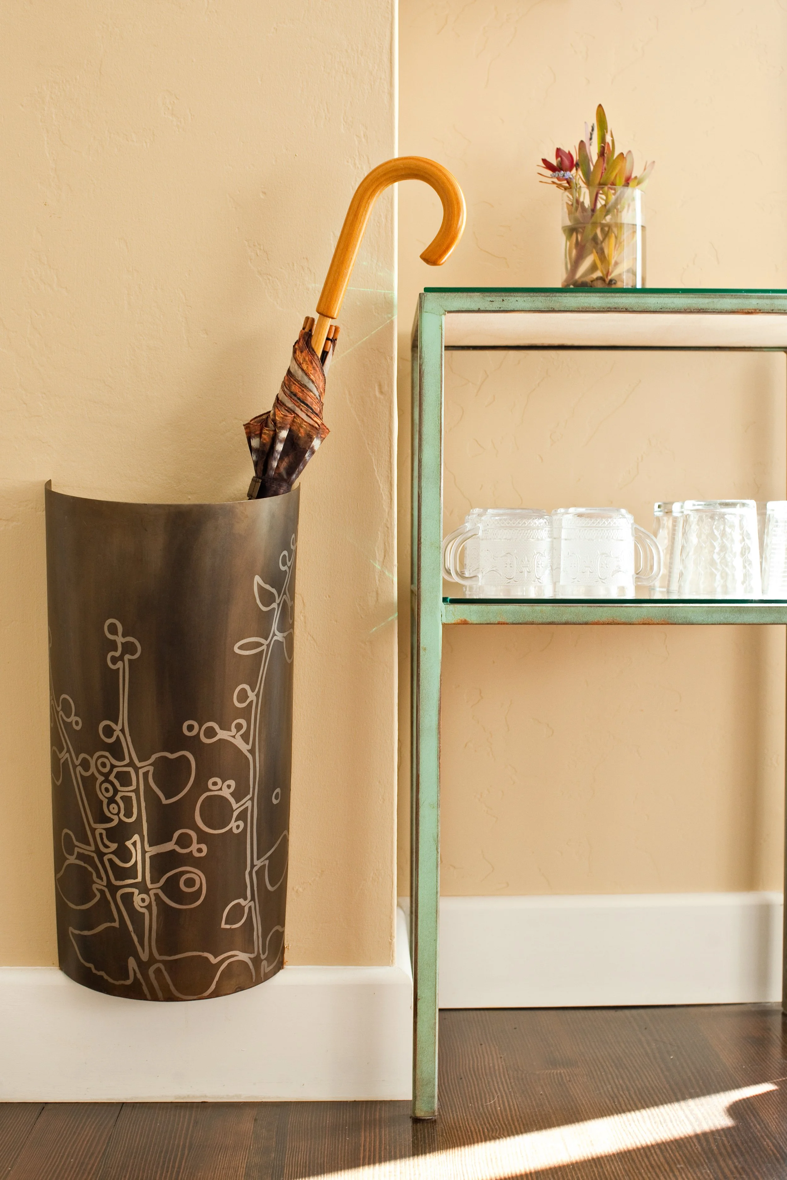 A black umbrella with a tree branch pattern leaning against a beige wall, and a glass shelf with a small glass container holding a plant with pink and green leaves. The shelf is supported by a light blue metal frame, with glass mugs underneath on a brown wooden floor.