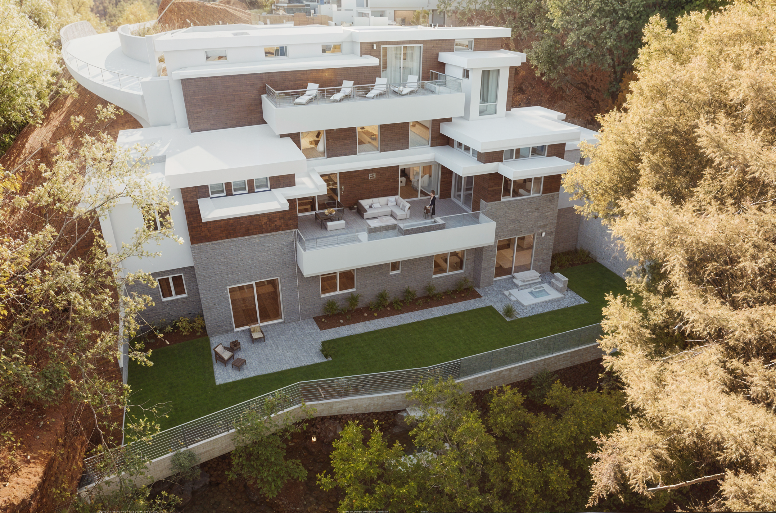 Aerial view of a modern multi-story residential building with white and brick exterior, multiple balconies with outdoor furniture, surrounded by trees and a landscaped backyard with patio area and grass.
