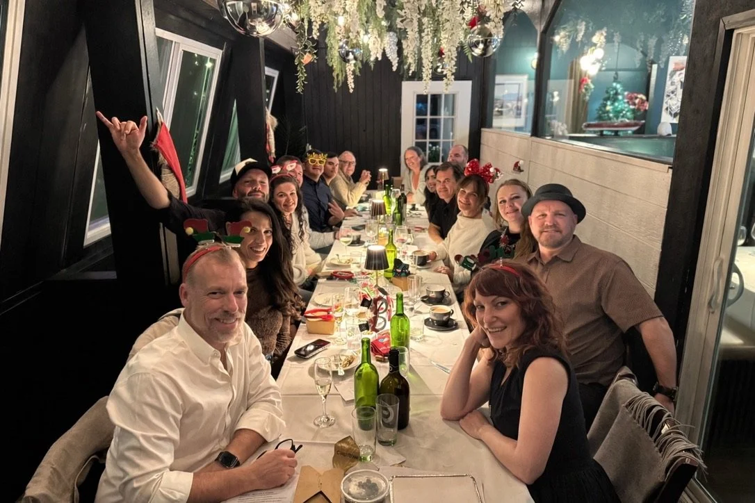 People gathered around a long table for a Christmas celebration, wearing festive accessories like reindeer antlers and Christmas crowns, in a decorated indoor setting.
