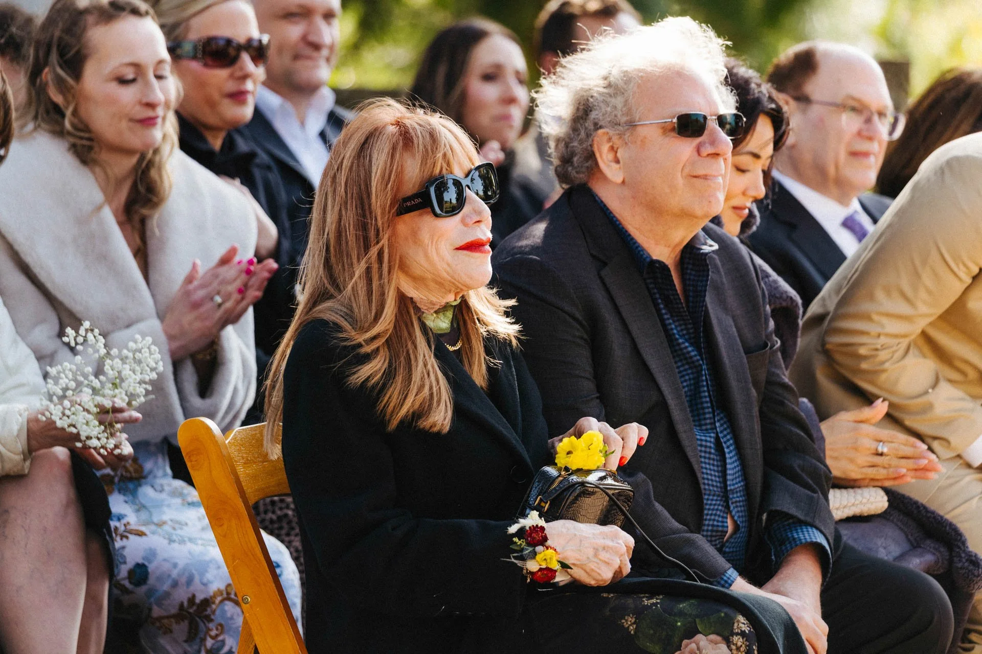 A group of older adults sitting outdoors during a sunny event, wearing sunglasses, with some holding flowers and dressed in semi-formal attire.
