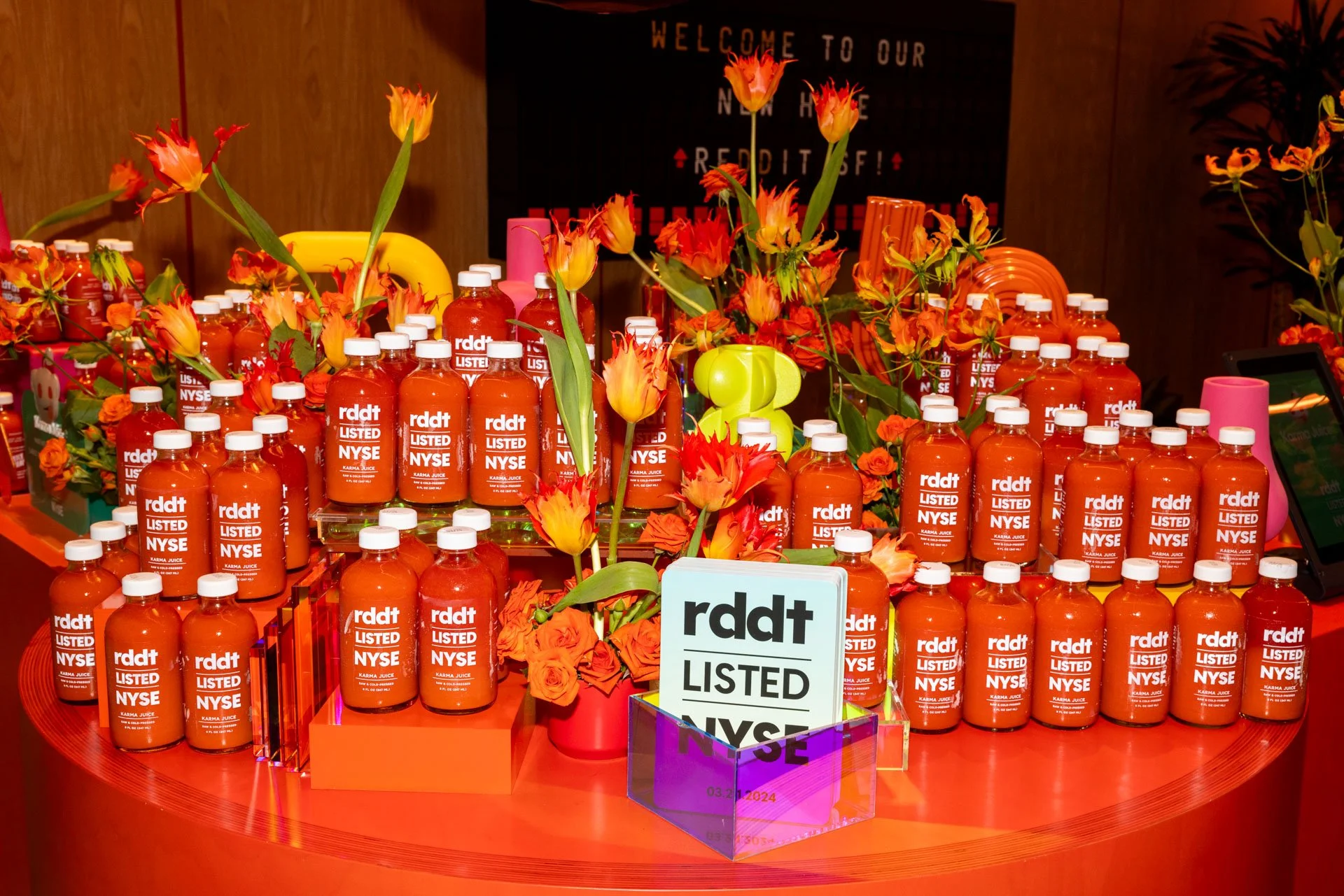Display of bottles labeled 'Rddt Listed NYSE,' arranged among orange and red flowers on a rounded table.