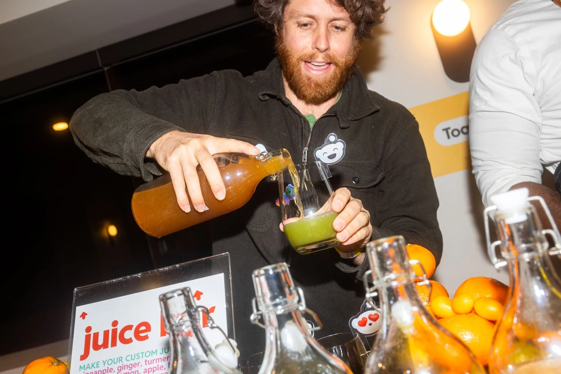 A man with red hair and a beard pours orange juice into a glass at a juice stand with oranges and juice bottles around.