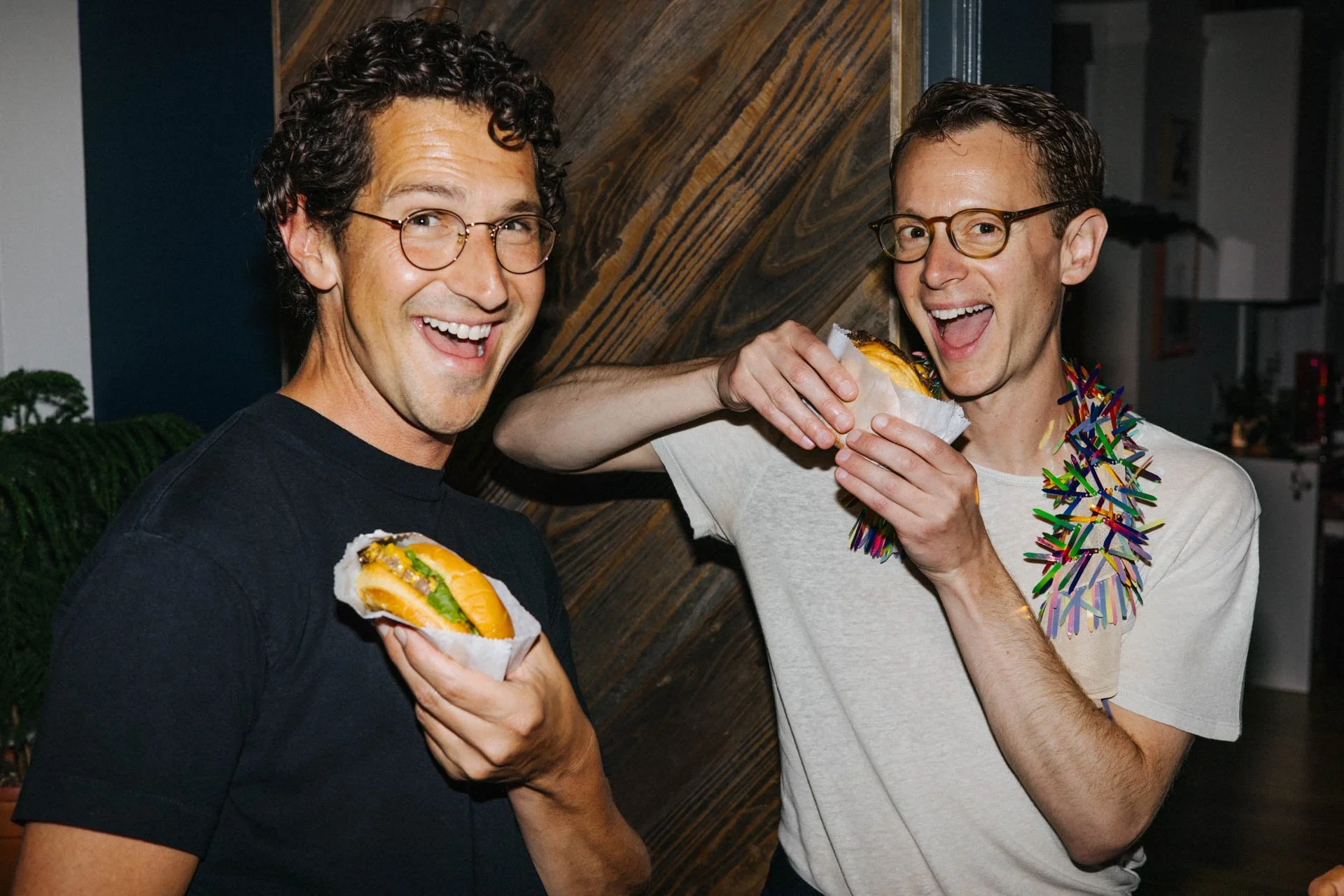 Two smiling men wearing glasses, holding hot dogs, with one also wearing a colorful party necklace, standing indoors.
