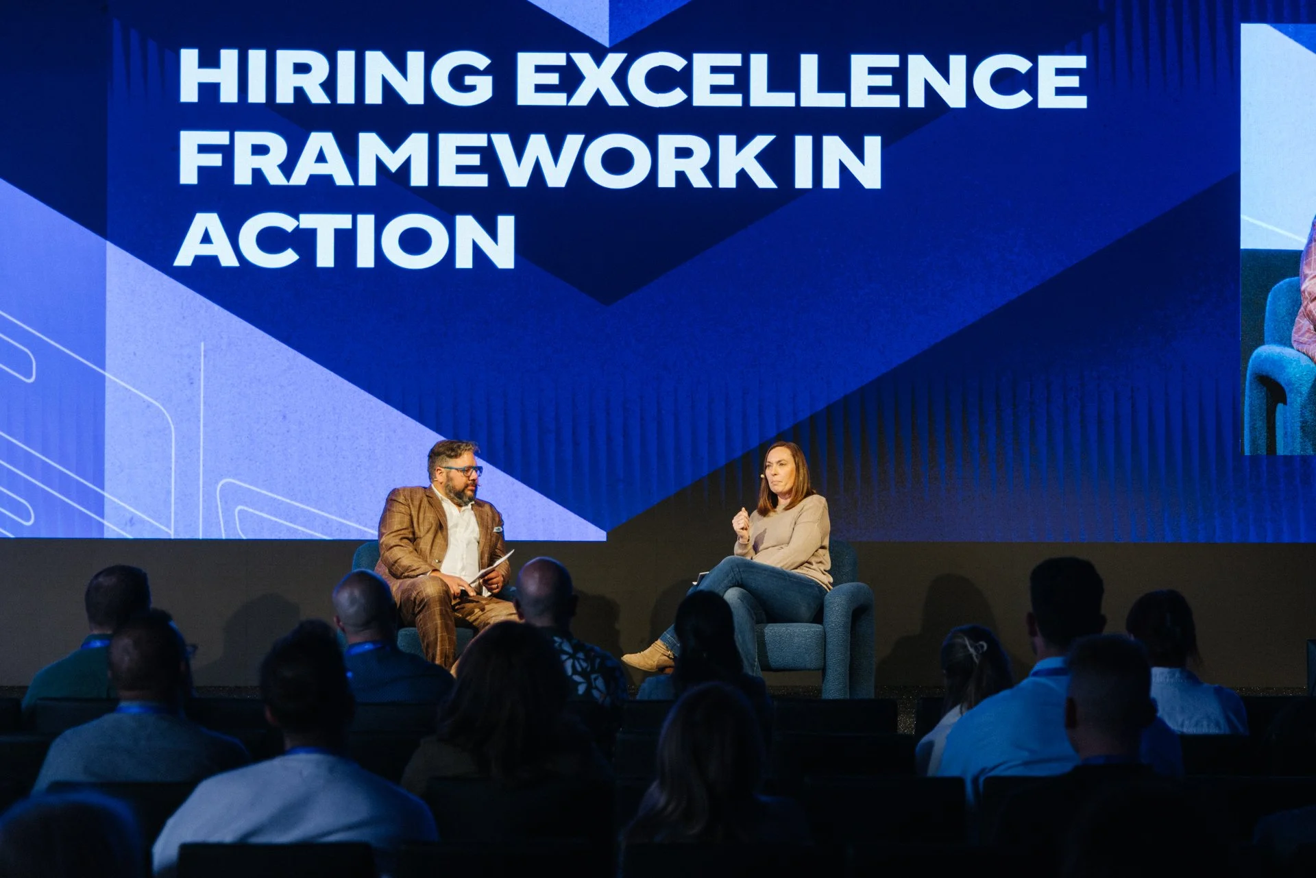 Two people sitting on stage in front of a large screen that reads "Hiring Excellence Framework in Action." One person is a man in a brown plaid suit holding a notepad, and the other is a woman in a beige sweater sitting with her legs crossed.