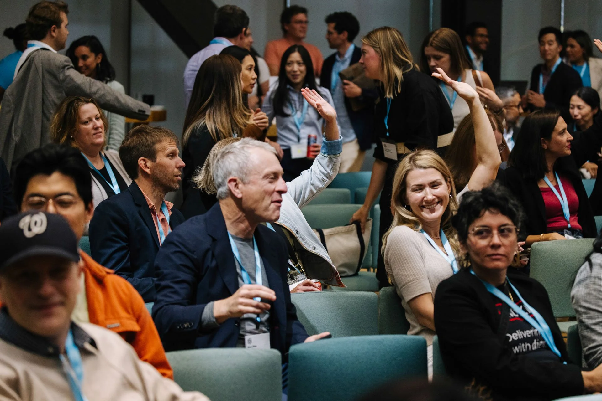 A diverse group of people attending a conference or seminar, with some standing and others seated, engaging with each other, smiling, and raising their hands.