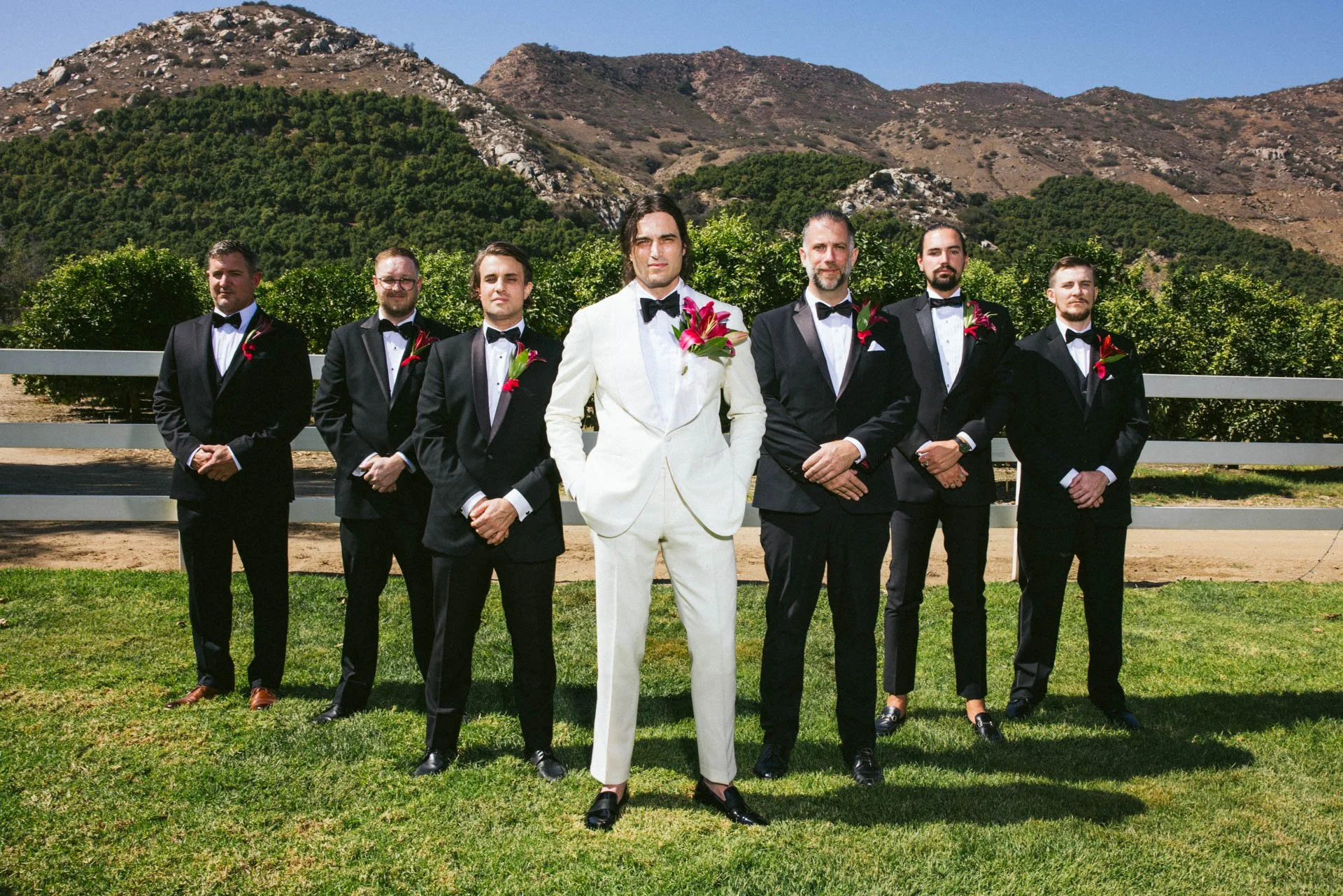 A groom in a white tuxedo and seven groomsmen in black tuxedos with bow ties and floral boutonnieres stand on a grassy area with a fence and mountain background.